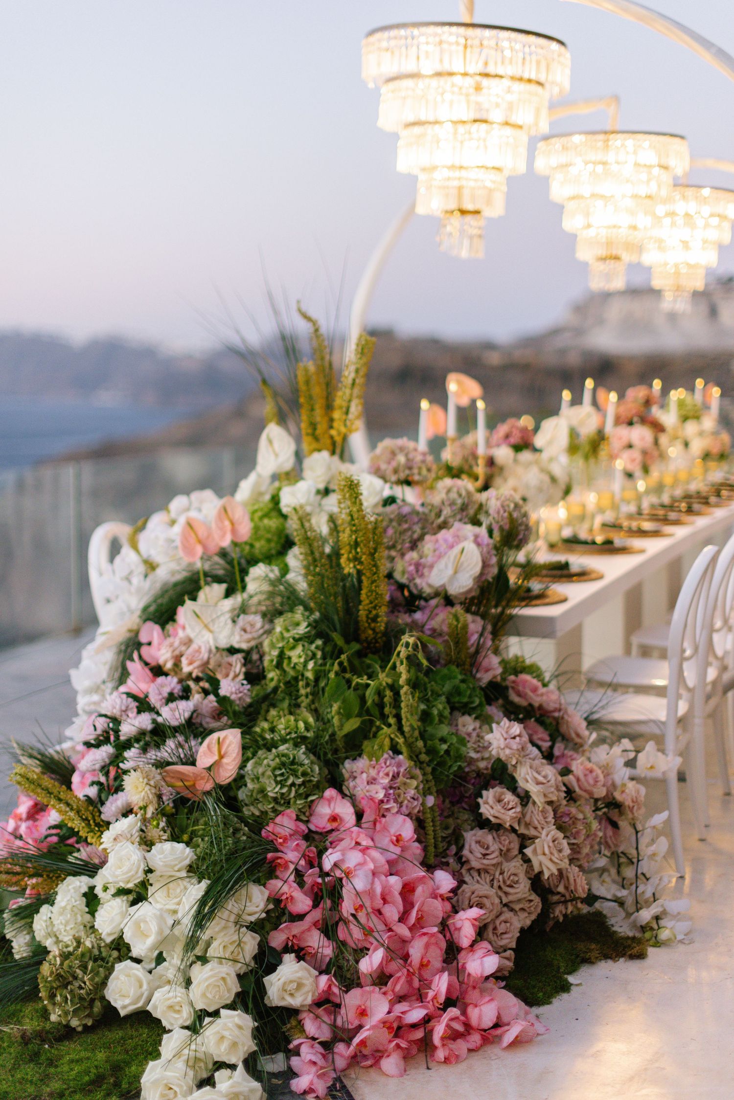 Elegant outdoor wedding reception table adorned with lush pink and white floral arrangements under crystal chandeliers.