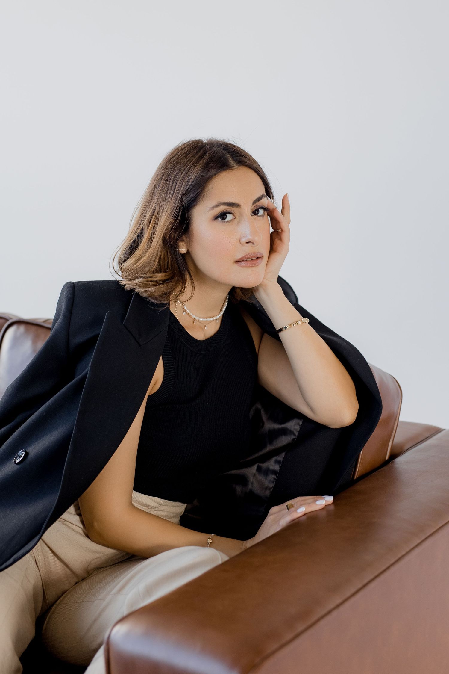 Woman in black blazer and beige pants reclines on brown leather couch.