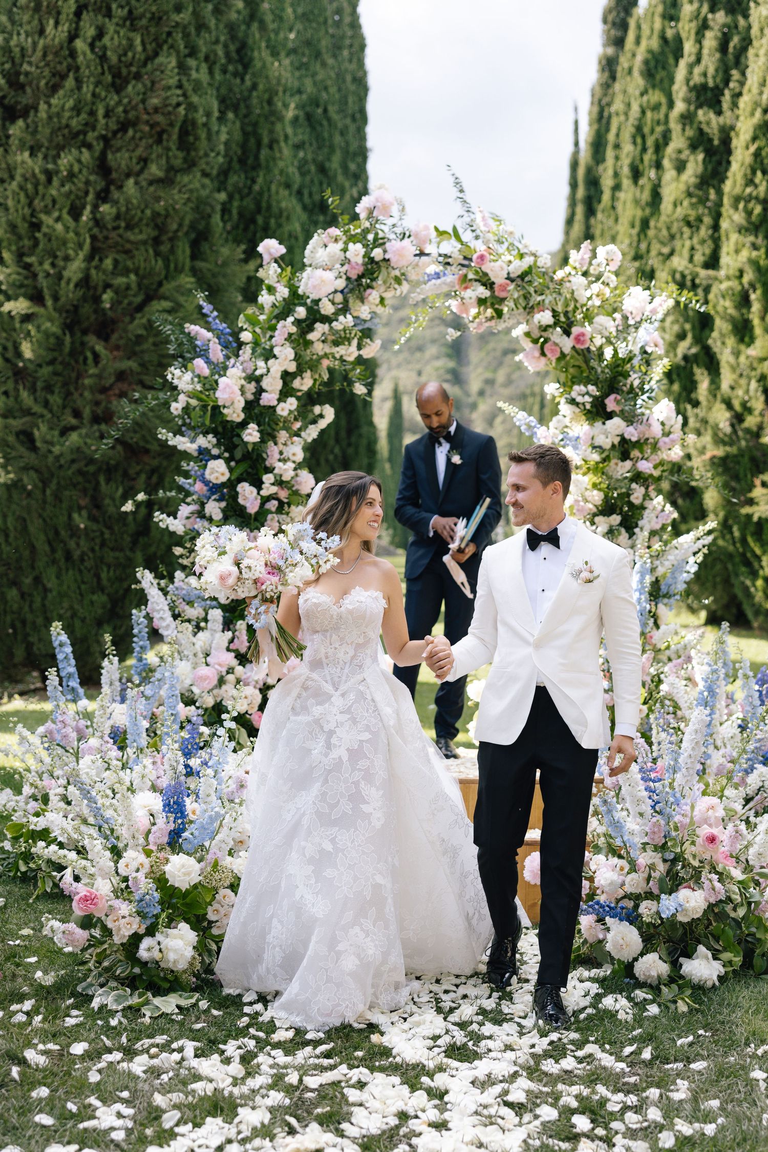 Bride and groom hold hands at outdoor wedding ceremony beneath floral arch of pink and blue blooms with cypress trees.