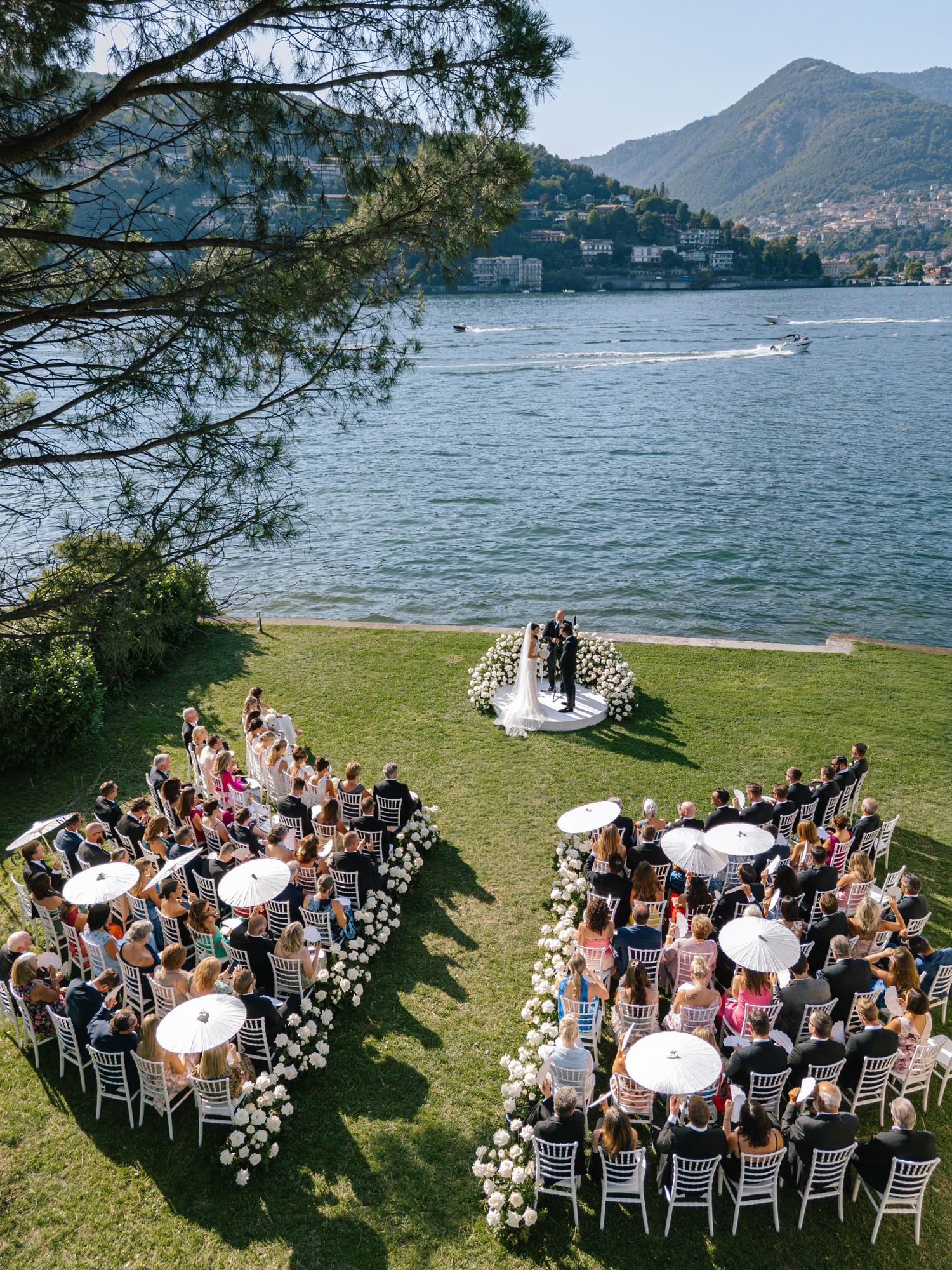 Aerial view of an outdoor wedding ceremony on a lush lawn beside a stunning Italian lake with mountains in the background.