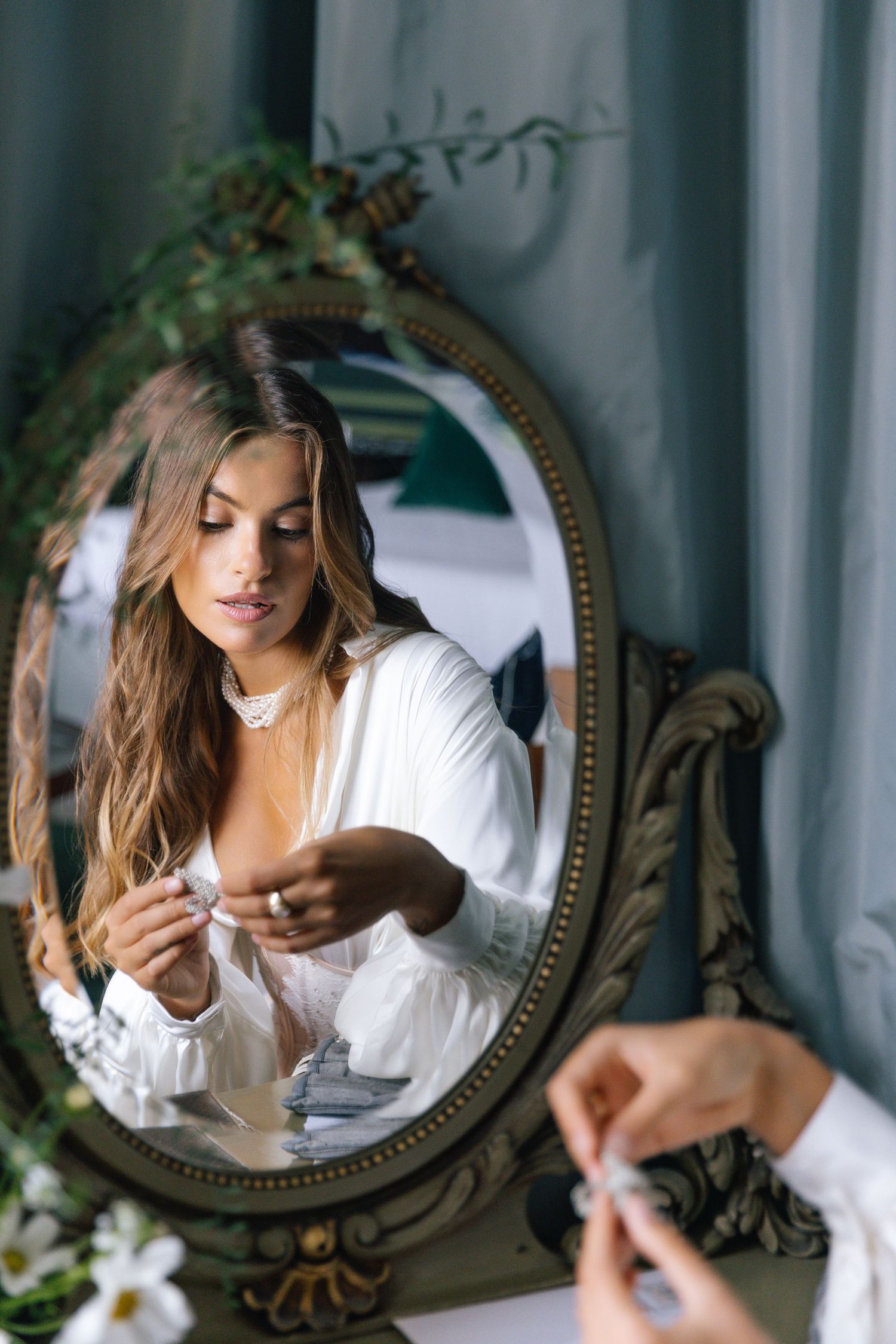 Reflection in ornate vintage mirror shows woman in white blouse adjusting jewelry near green foliage decor.
