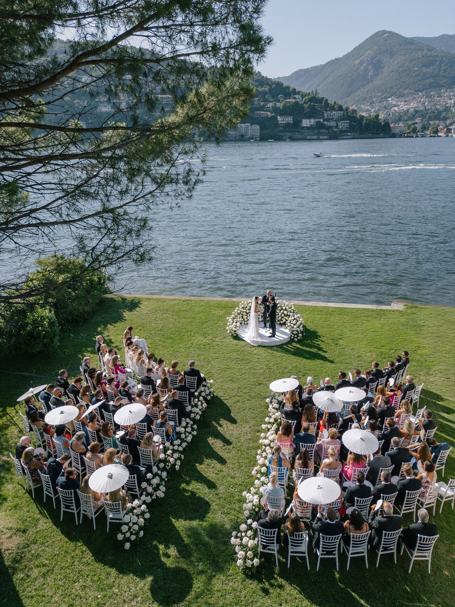Scenic lakeside wedding ceremony setup with mountain views and guests seated in white chairs by Lake Como, Italy.