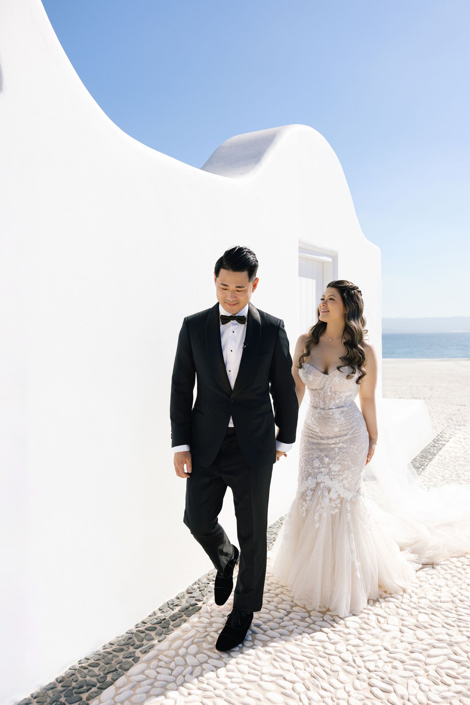 A couple in wedding attire walks along a white-walled Mediterranean path with ocean views on their wedding day.