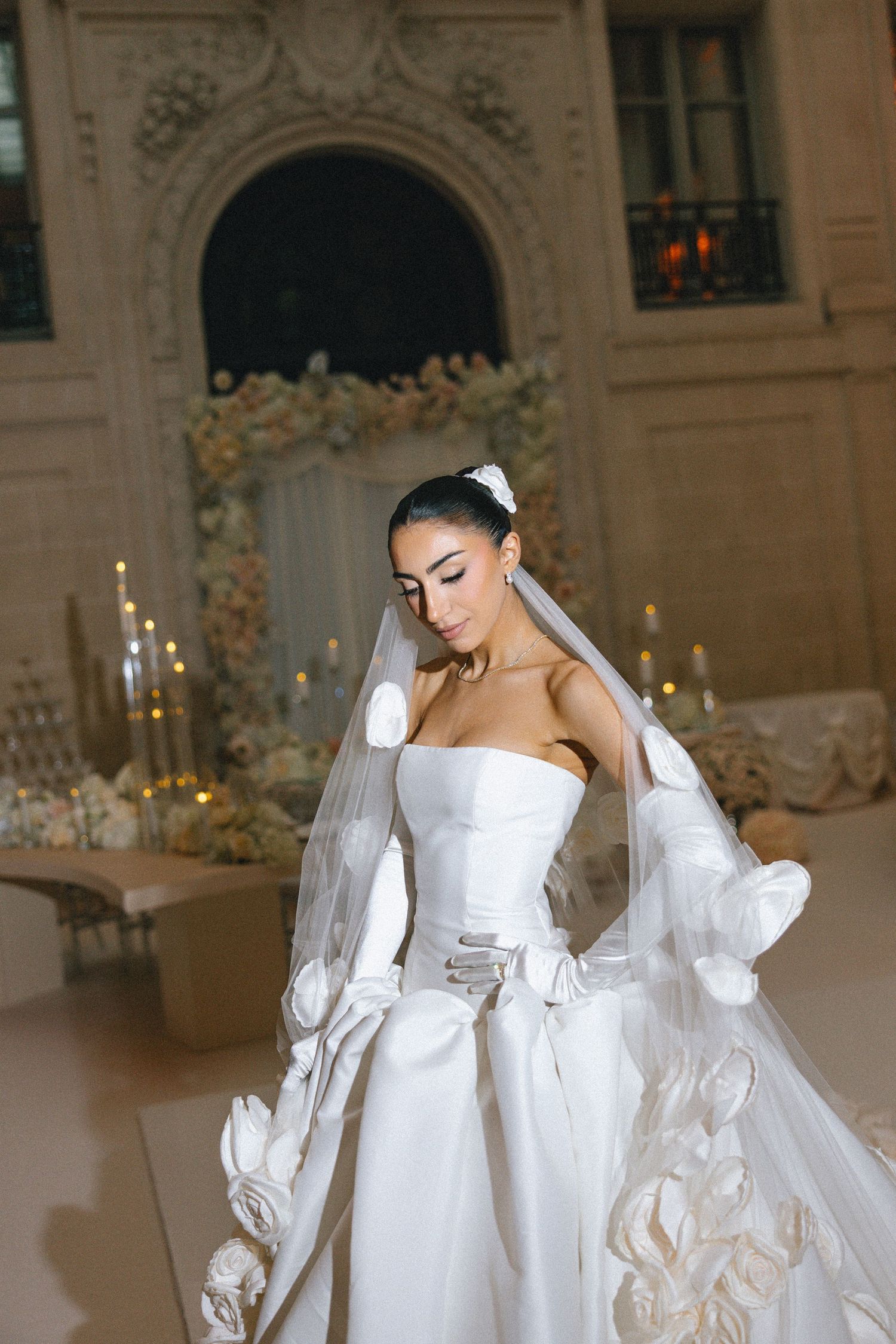 A bride in an elegant white long-sleeved wedding gown poses in an ornate ballroom with glowing candelabras.