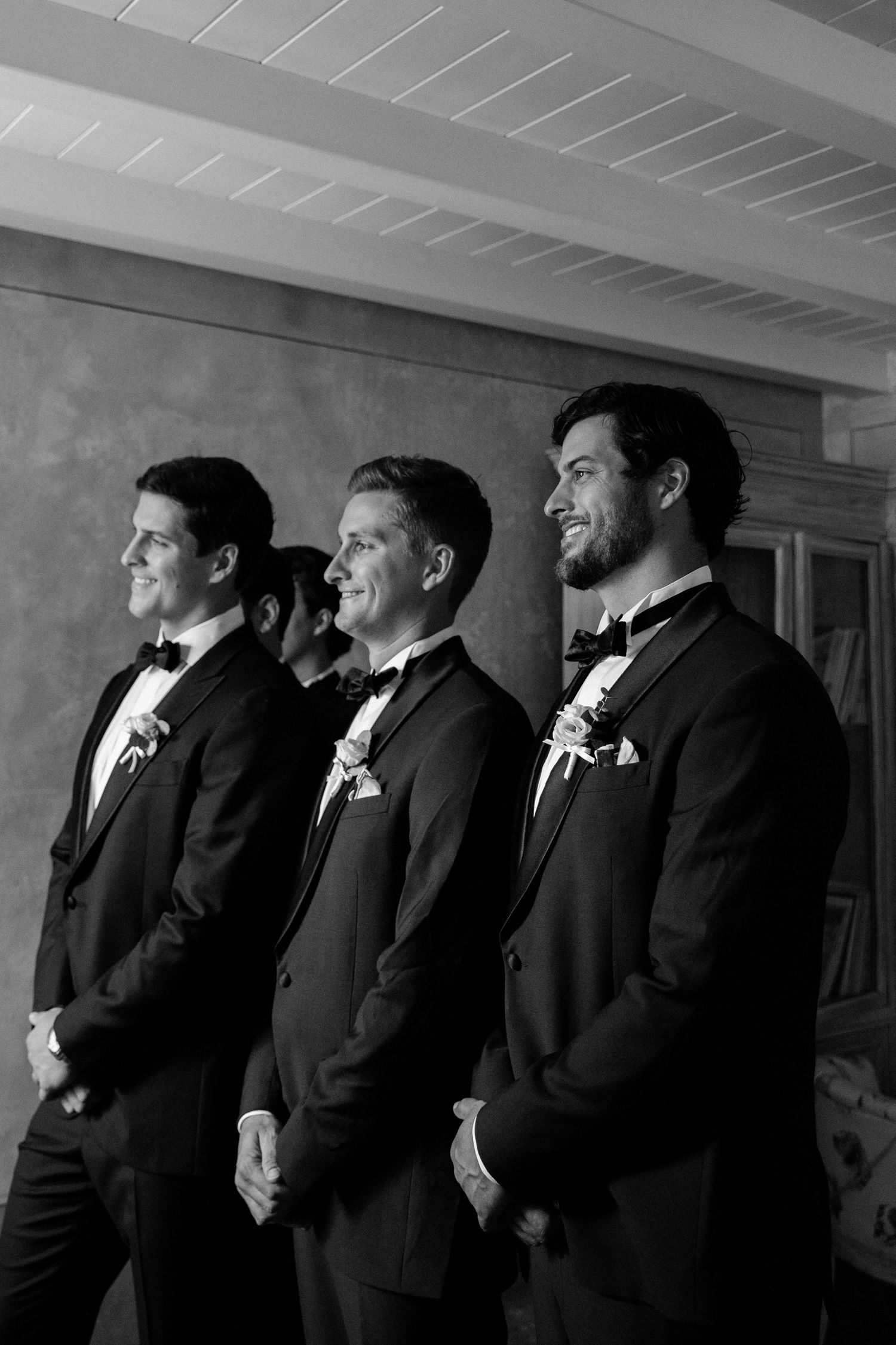 Three groomsmen in black tuxedos with boutonnieres smile during a wedding ceremony in a black and white photo.