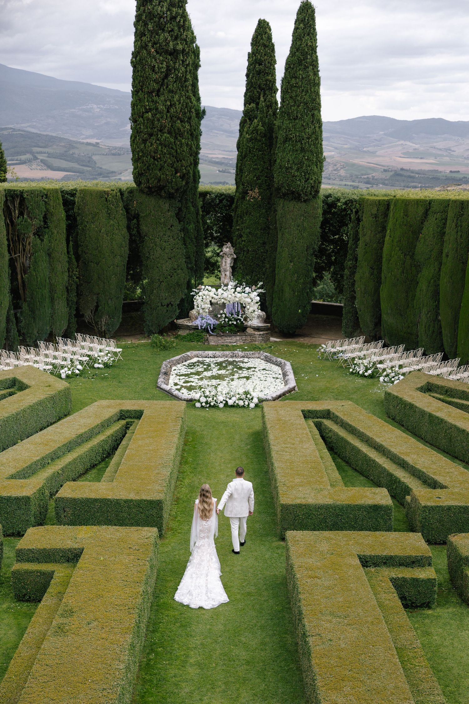 Bride and groom walk through elegant Italian garden with manicured hedges, white floral altar, and cypress trees at outdoor wedding.