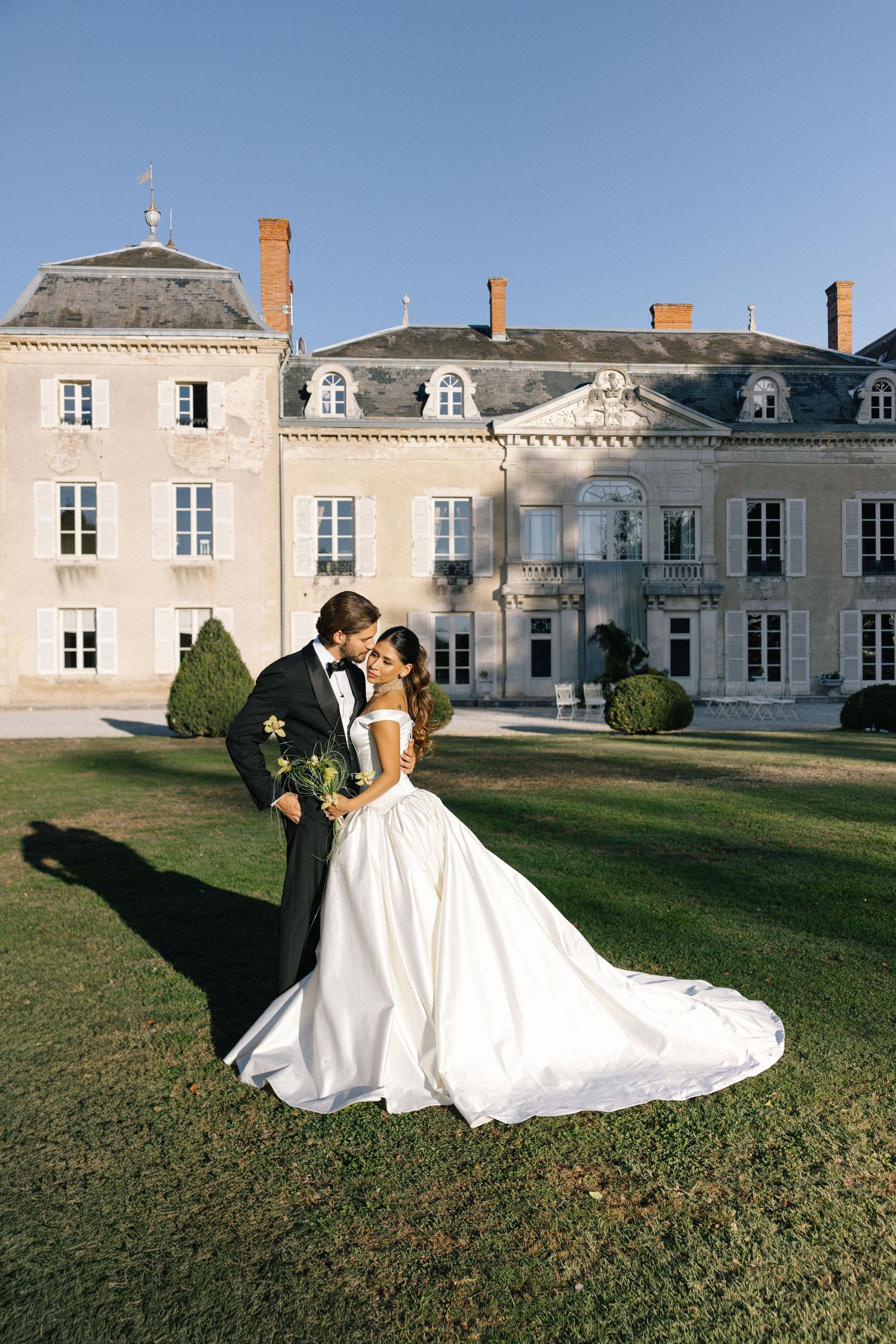 Bride and groom share a romantic kiss on green lawn before elegant French château on a sunny wedding day.