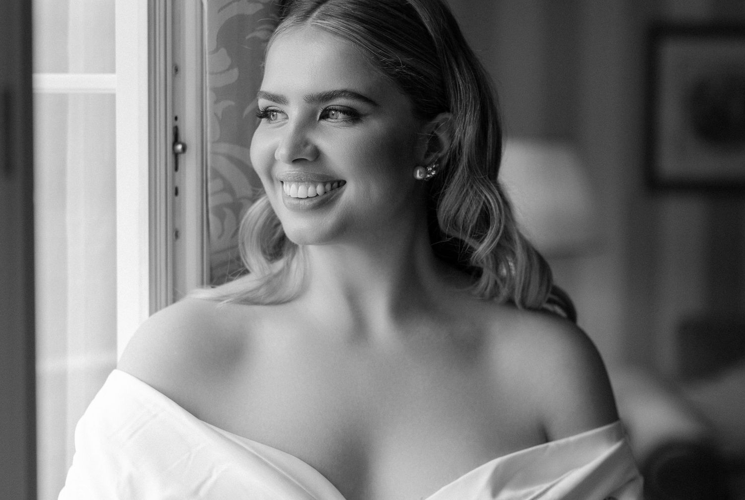 Black and white portrait of a smiling bride in an off-shoulder gown looking towards window light.
