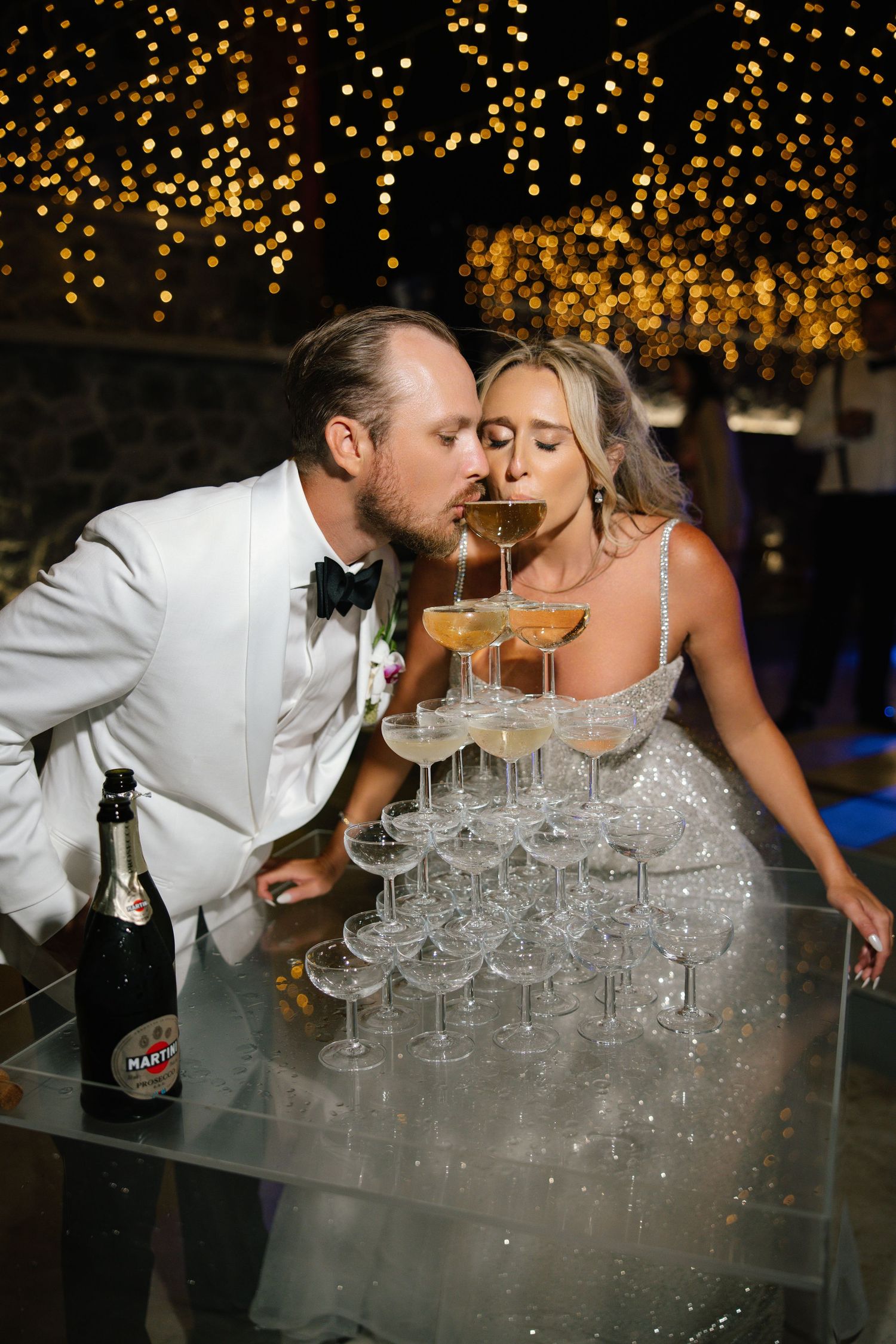 Bride and groom kissing over a champagne tower at their wedding reception with golden bokeh lights in the background.