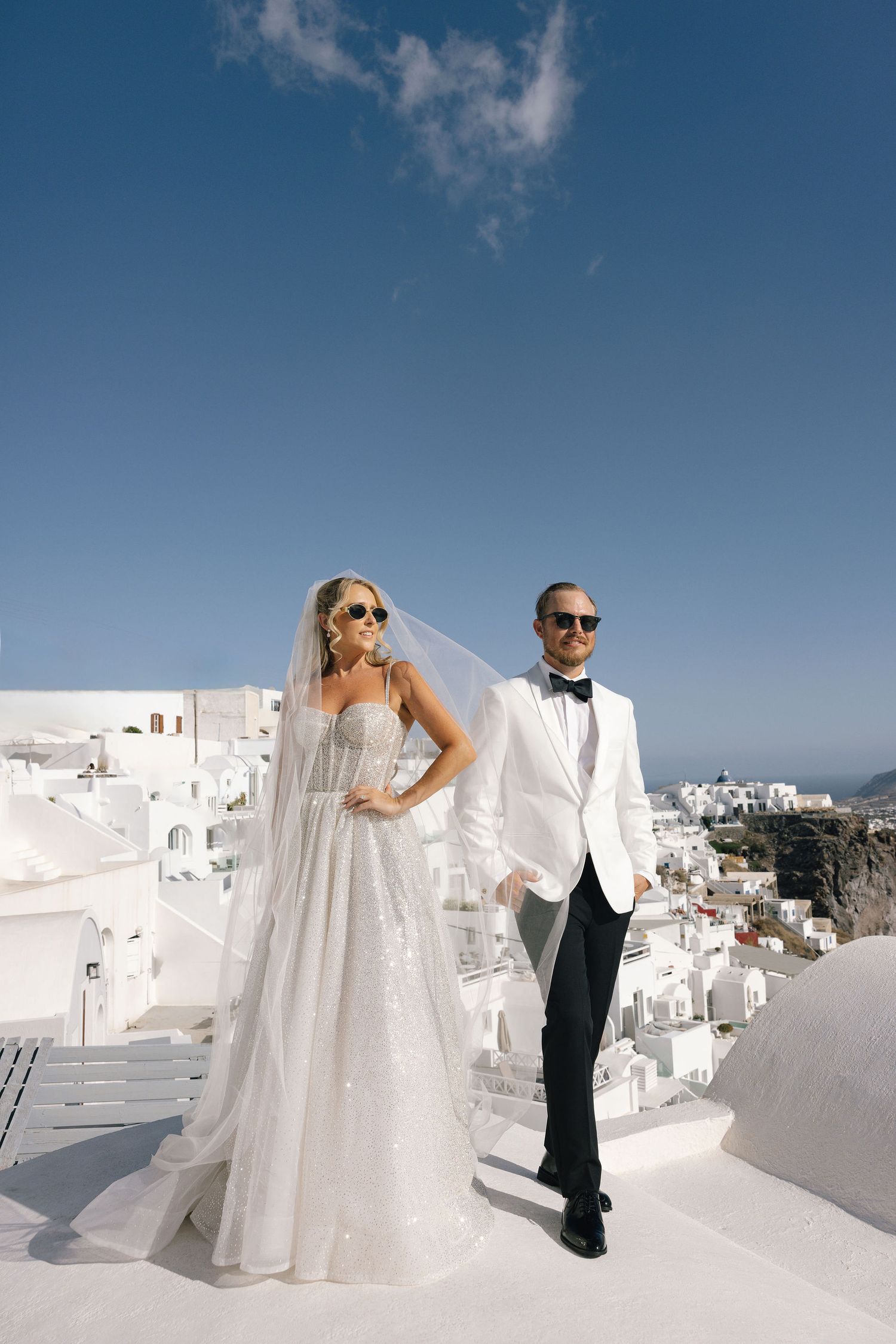 Bride and groom in sunglasses pose on white rooftop in Santorini, Greece, with stunning blue sky backdrop.