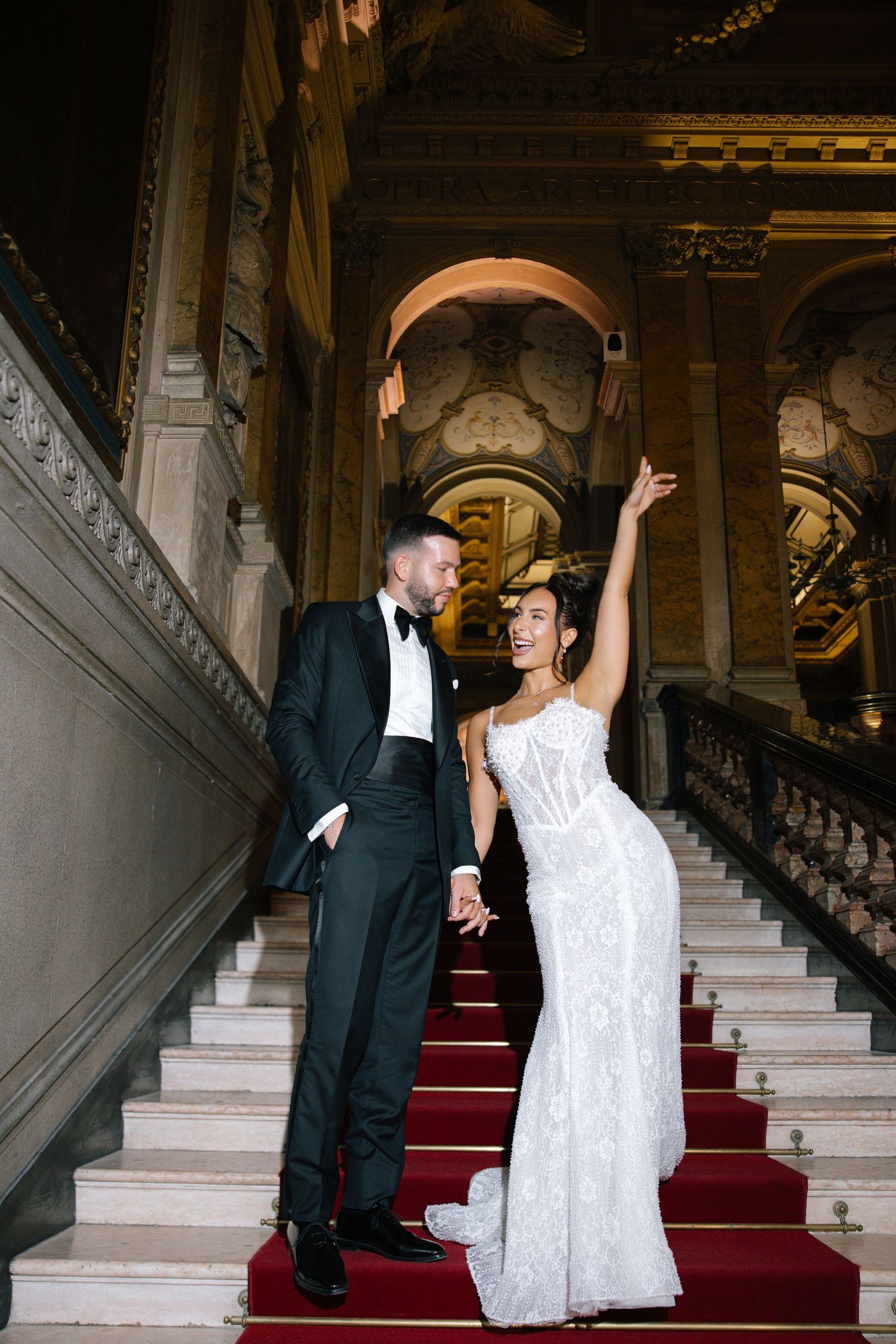 Bride and groom holding hands on grand staircase with red carpet in elegant venue.