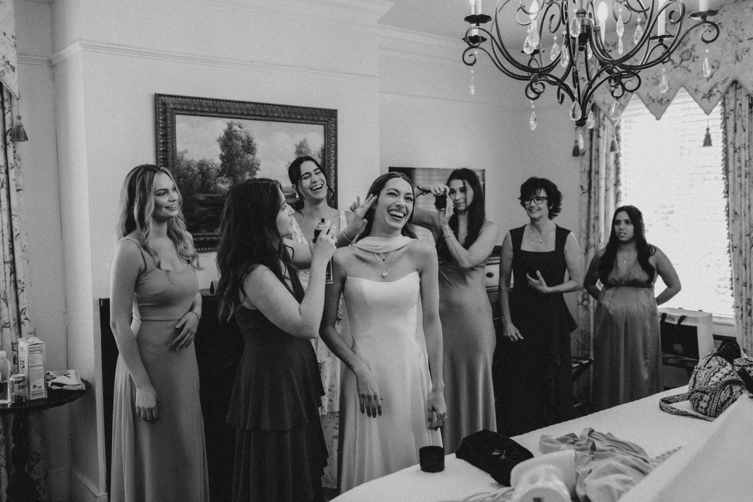 A bride stands in the middle of a group of women, including bridesmaids and mothers, as they adjust her hair and smile