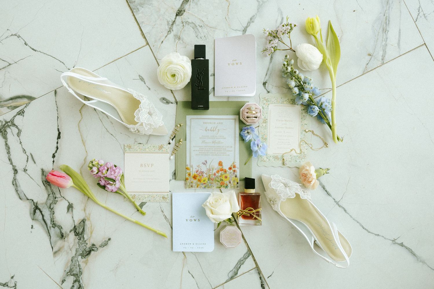 A flat lay photograph of wedding invitations, flowers, rings, perfume, and shoes