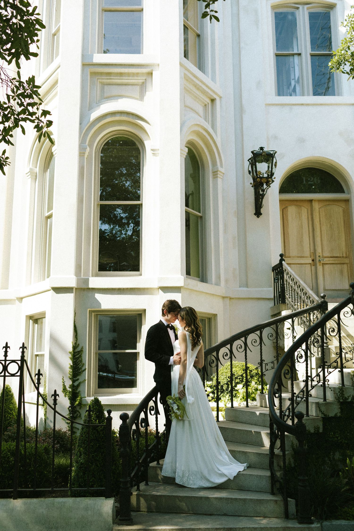 A wide-shot image of a bride and groom standing on a curved staircase outside a white mansion, touching their foreheads together