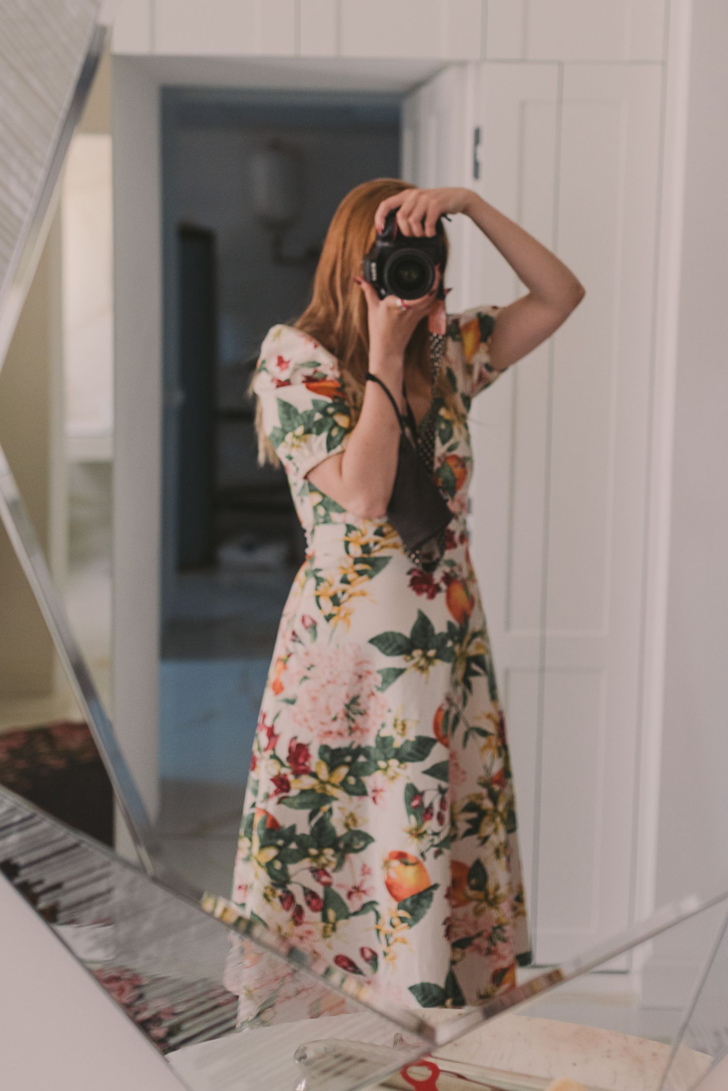 Woman wearing a floral dress standing in a white hallway taking mirror selfies.