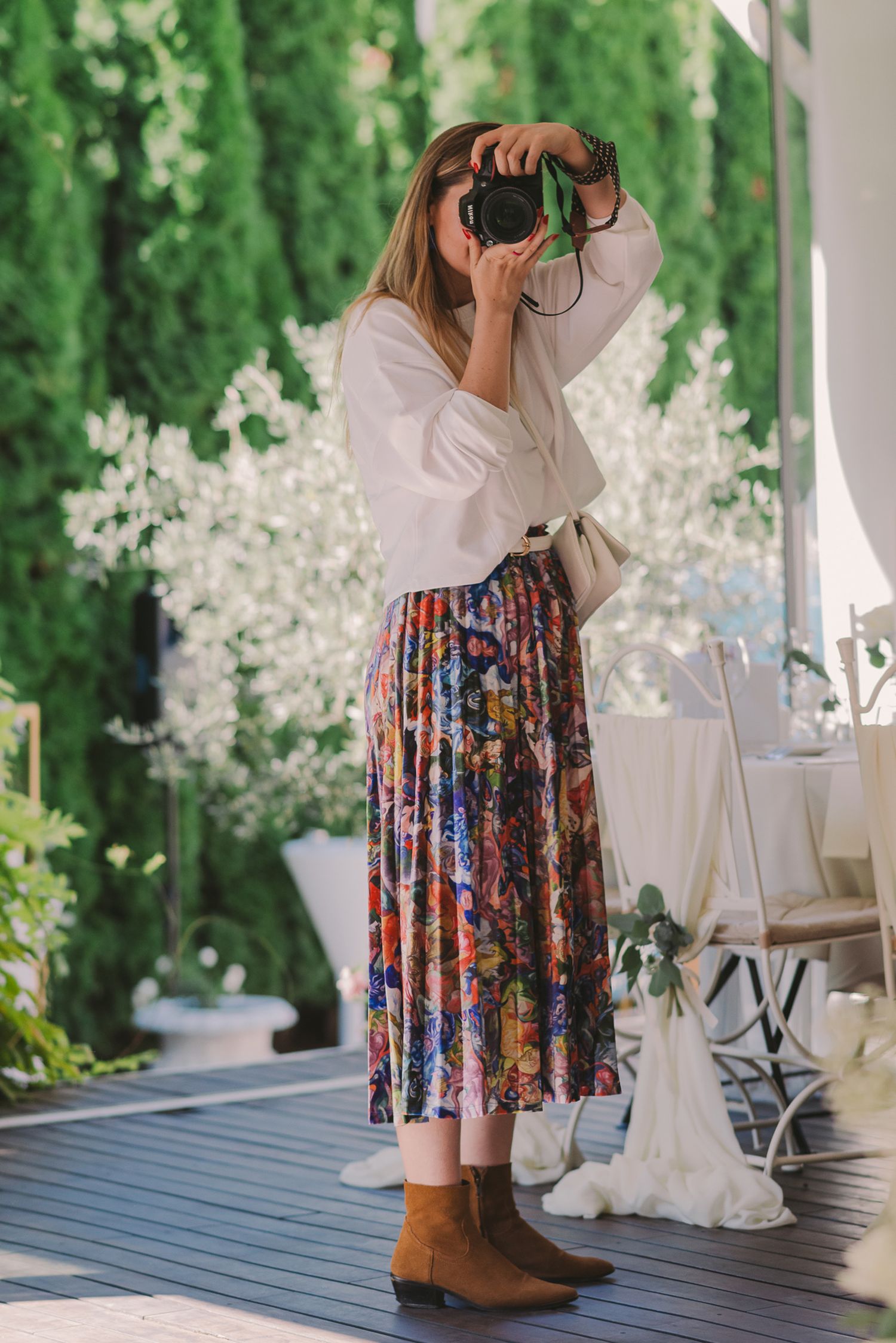 A person wearing a white top and floral skirt takes photos with a camera on an outdoor porch with greenery in background.