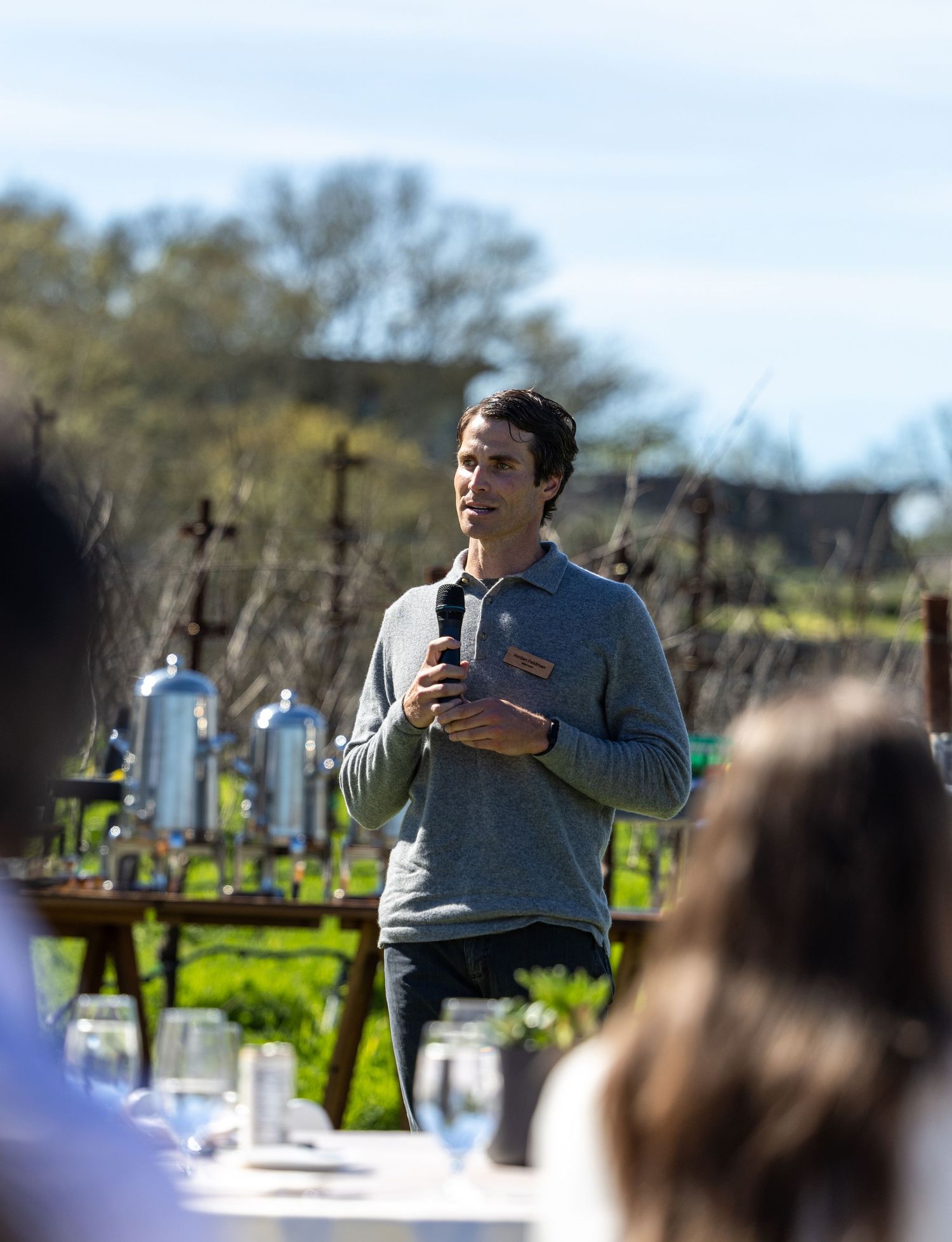 Man in gray polo speaks to outdoor vineyard audience while holding a microphone at a winery event.