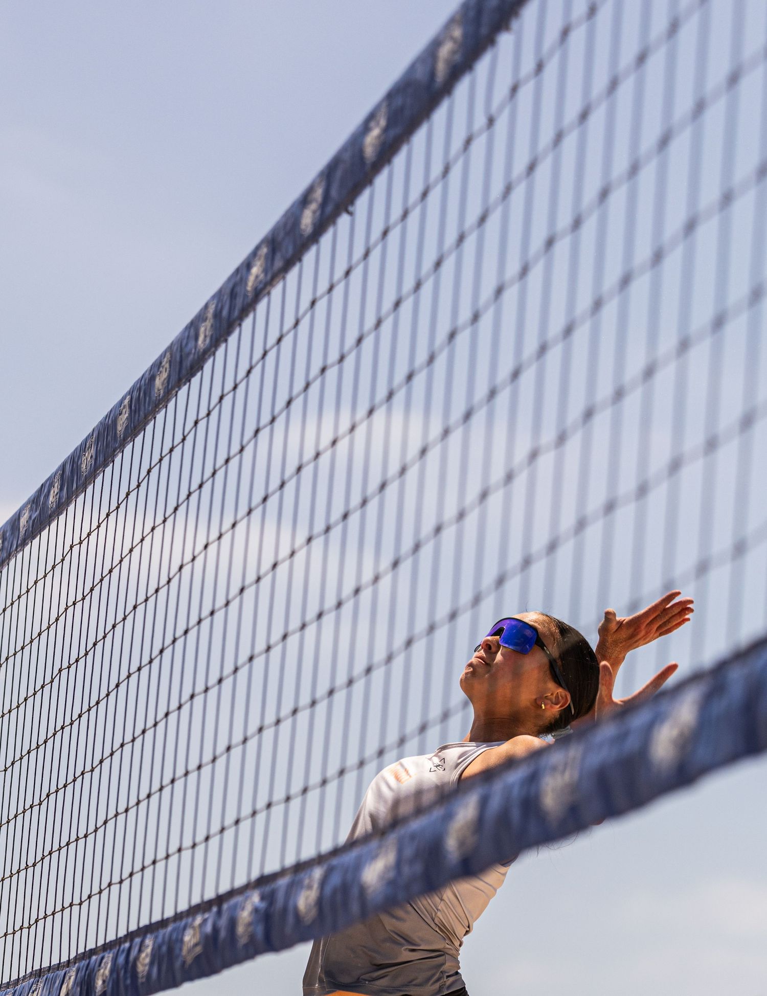 A beach volleyball player in sunglasses jumps to spike the ball at the net against a blue sky.