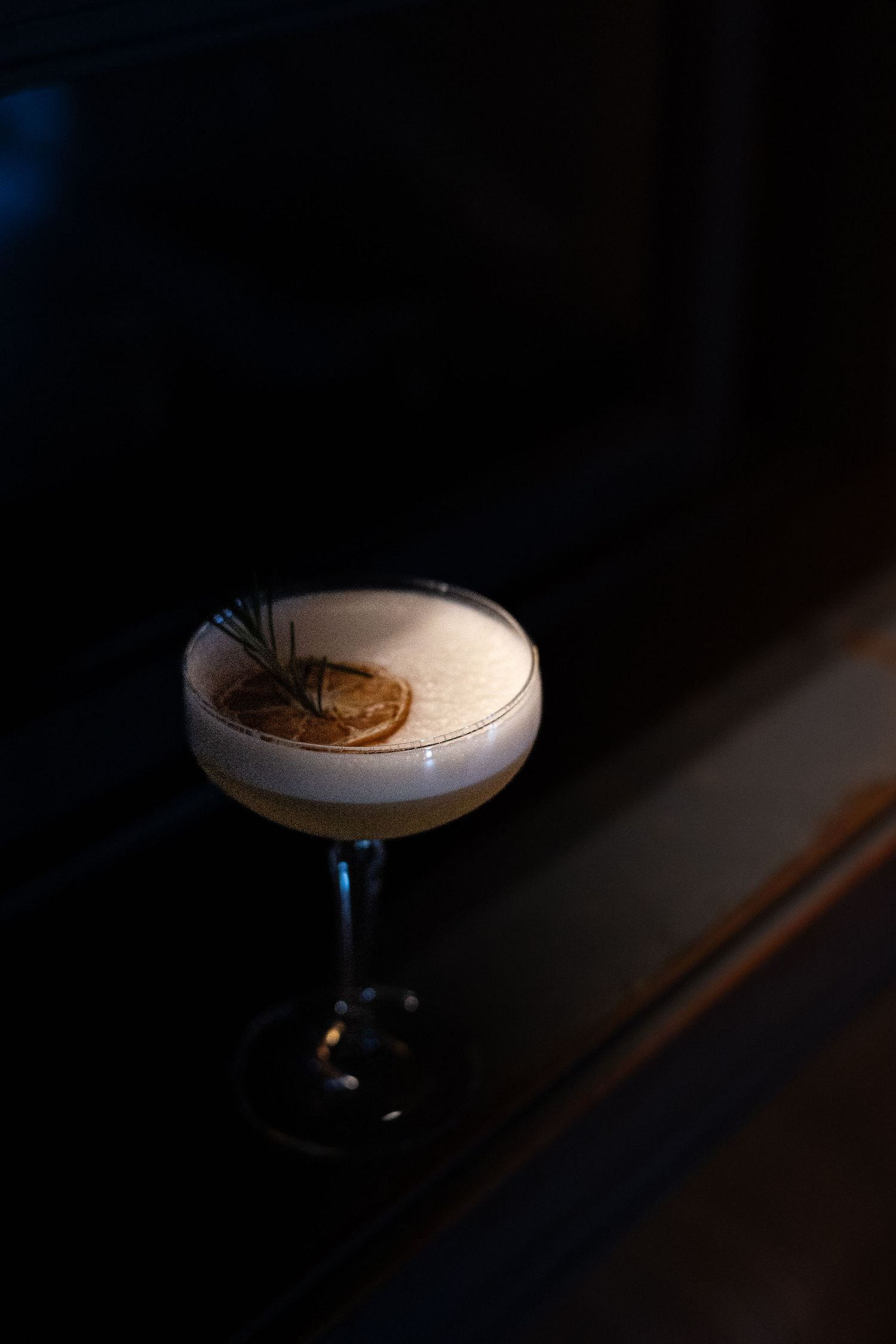 Elegant cocktail in a coupe glass with frothy top, garnished with a dried citrus slice and rosemary sprig in dark ambiance.