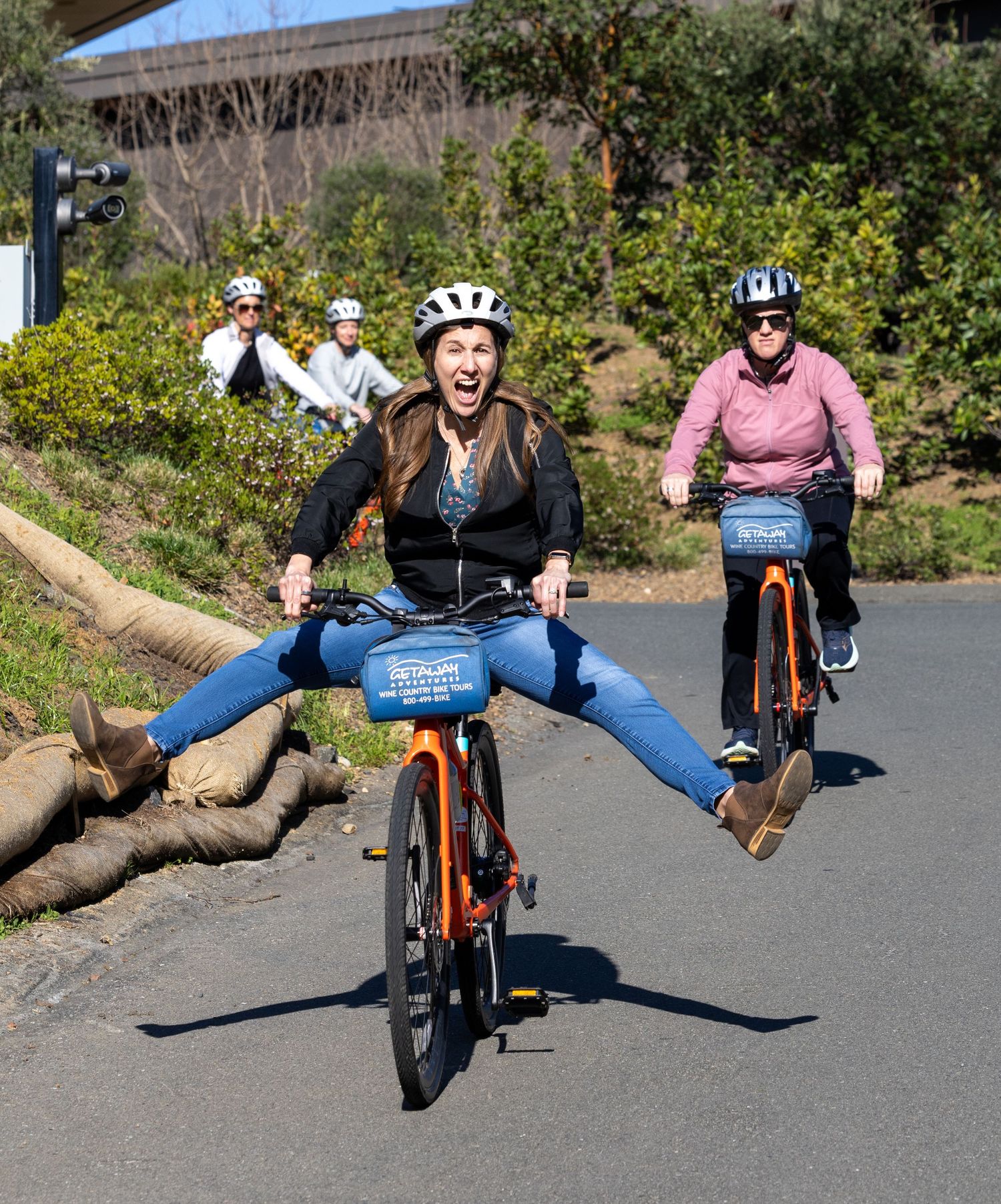 Woman joyfully riding a bike with legs out on a sunny path, followed by other cyclists wearing helmets.