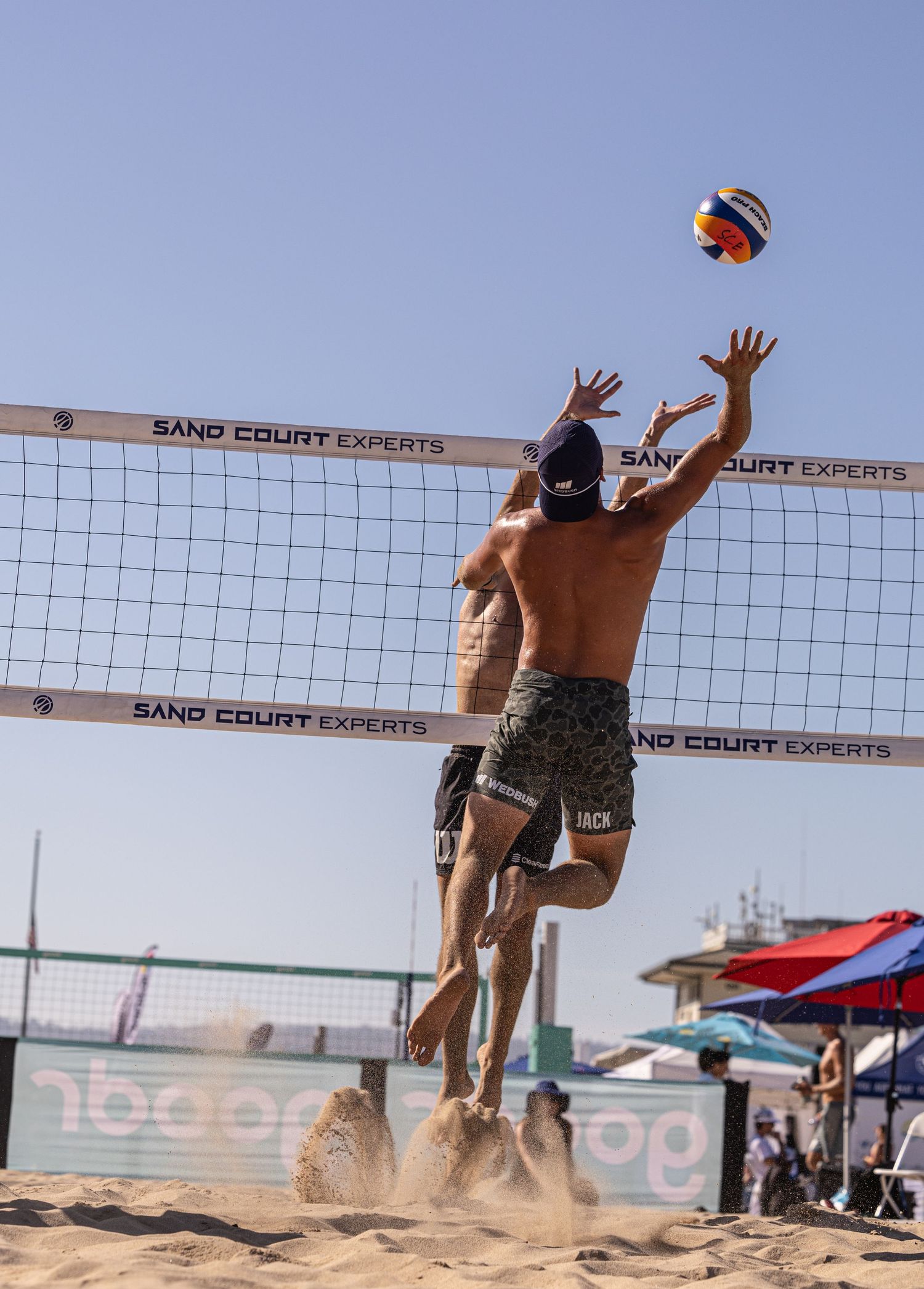 Two men playing beach volleyball, one jumping to spike the ball over the net at a Sand Court Experts tournament.