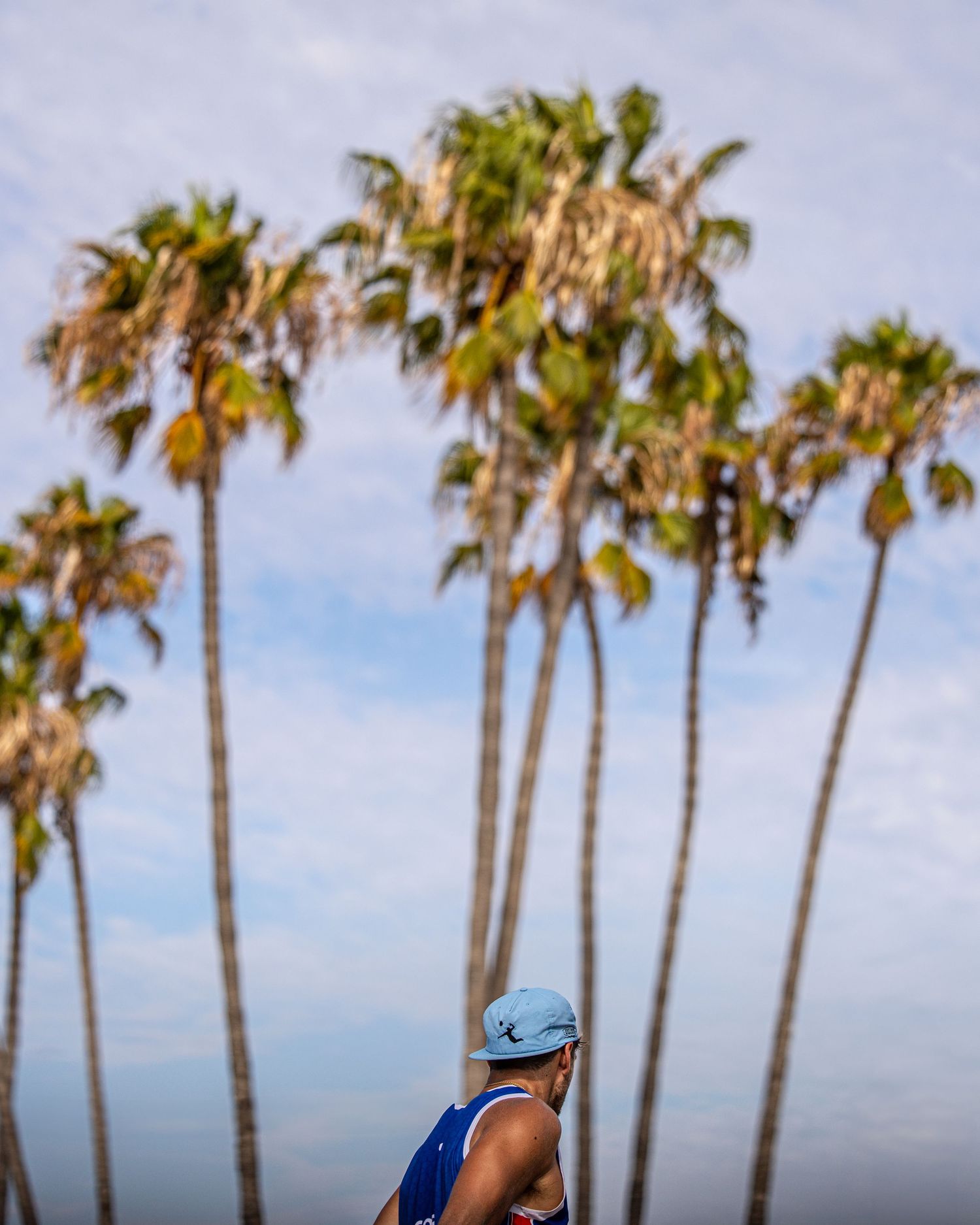 Athlete in blue cap and tank top stands before tall palm trees against a partly cloudy sky.