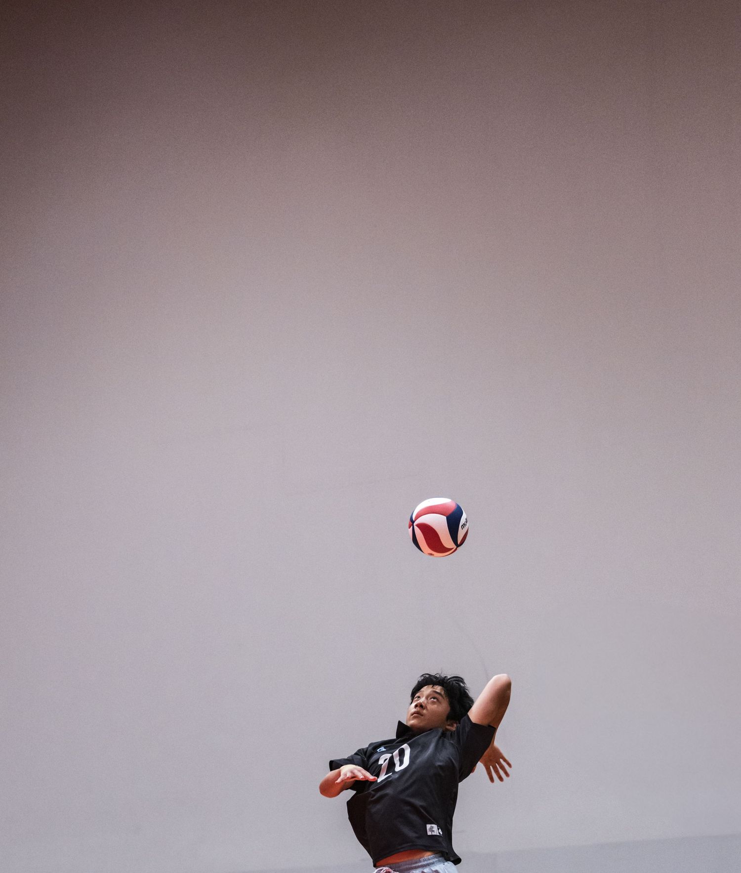 A young volleyball player in a black jersey leaps to serve a red, white, and blue volleyball indoors.
