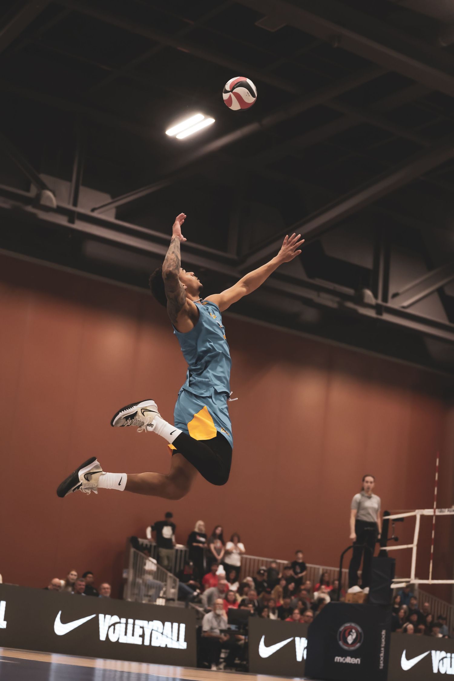 Male volleyball player leaping high to spike ball during indoor match, Nike branding visible on court sidelines.