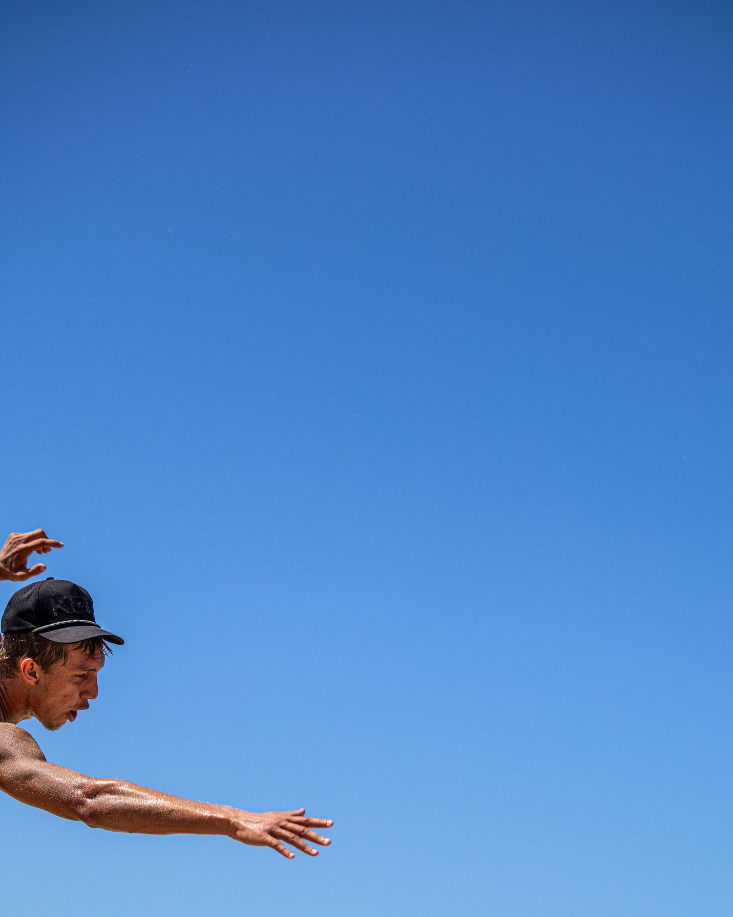 A man in a black cap leaps against a clear blue sky, arms outstretched in a dynamic mid-air pose.