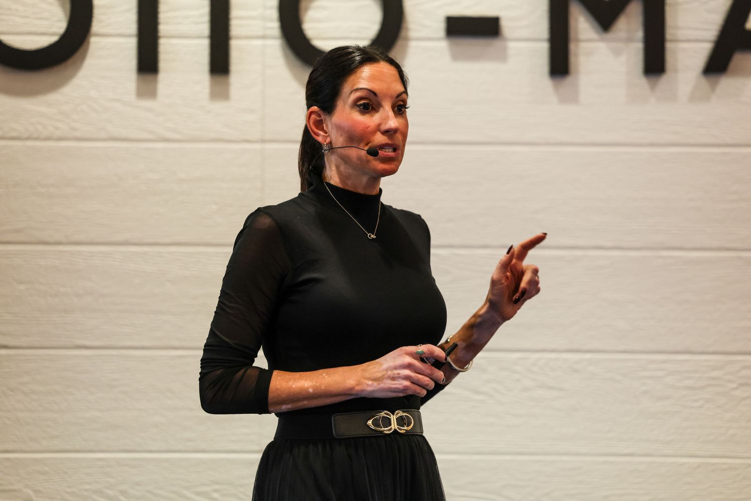 Woman in black outfit speaking at an event, gesturing with one finger raised, microphone headset on.