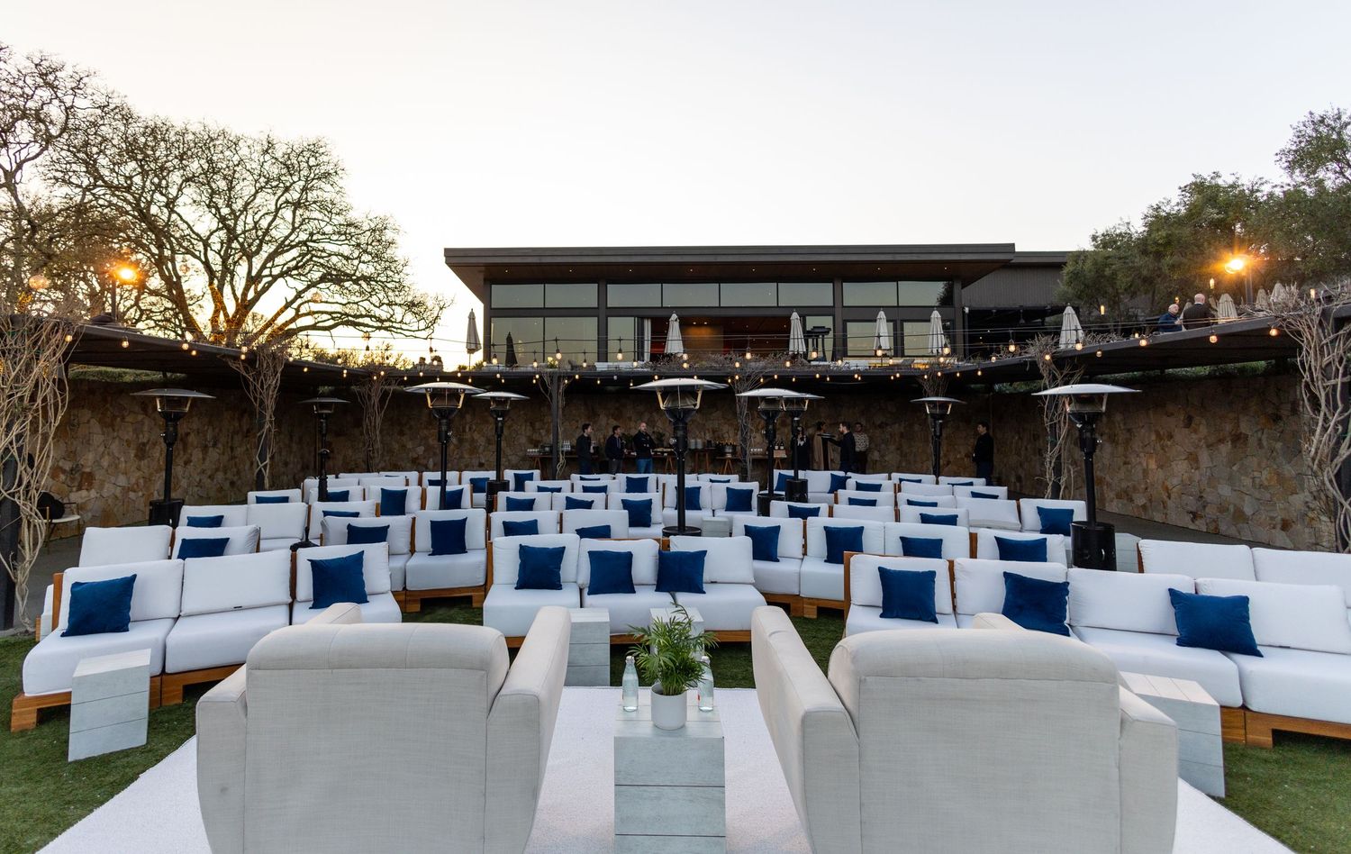 Elegant outdoor lounge area with white sofas, navy pillows, and patio heaters set up for an evening event.