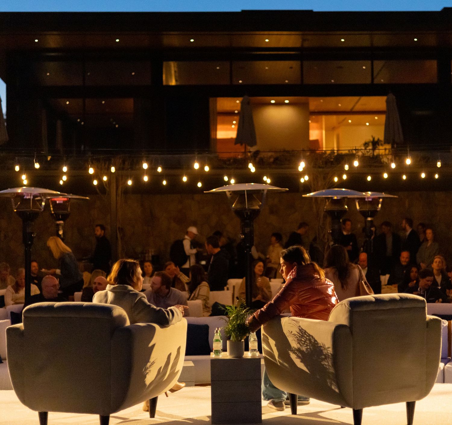 Two people in armchairs at an outdoor evening networking event with string lights.