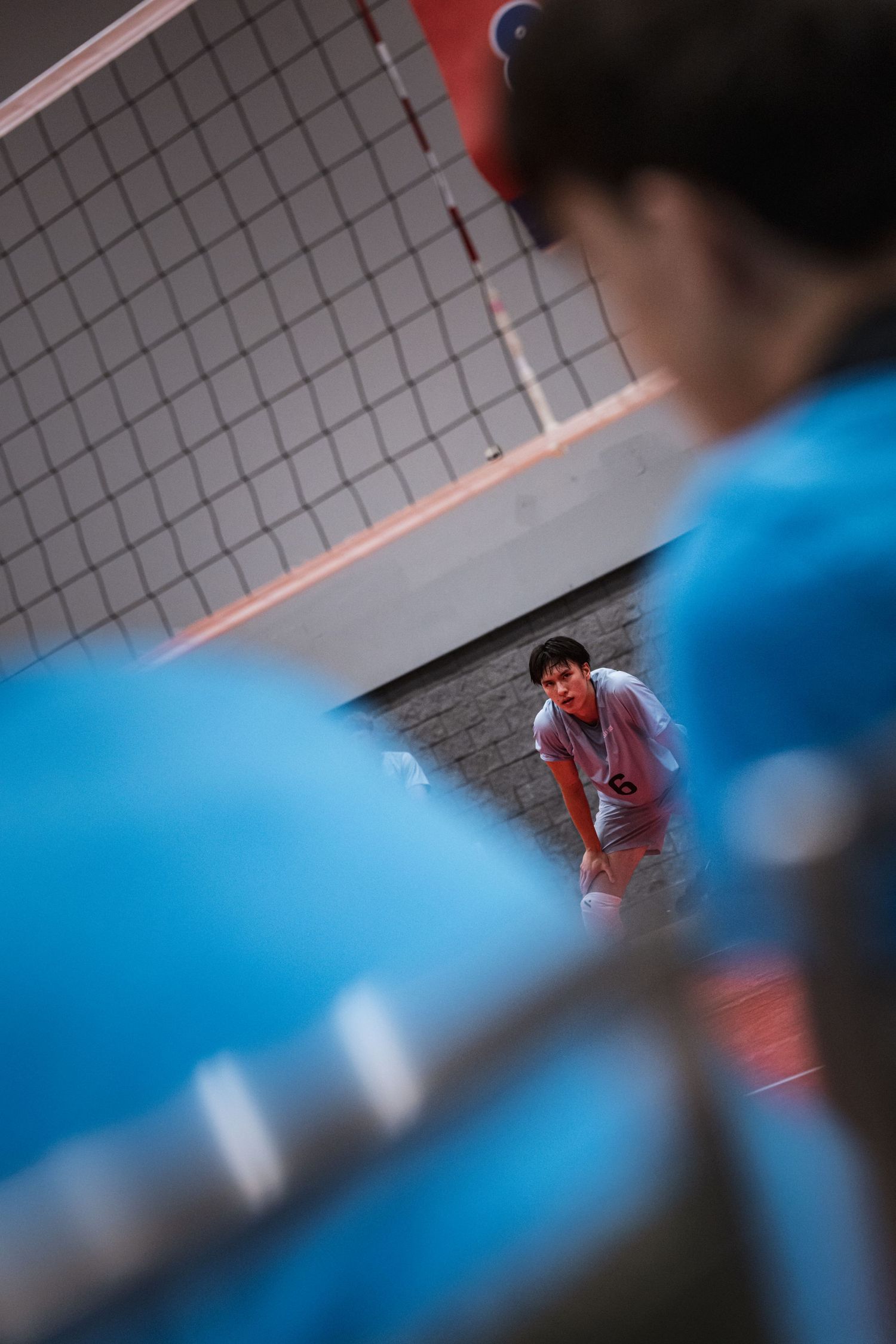 Volleyball player in blue jersey crouches ready to receive serve on indoor court.