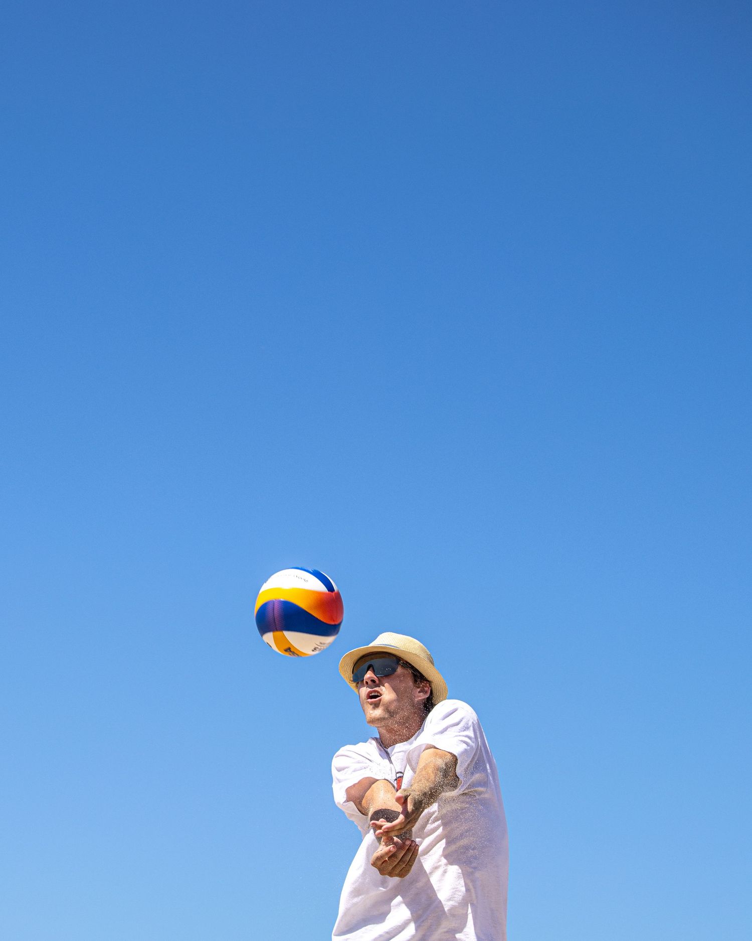 Man in white shirt and straw hat playing beach volleyball, bumping a colorful ball against a clear blue sky.
