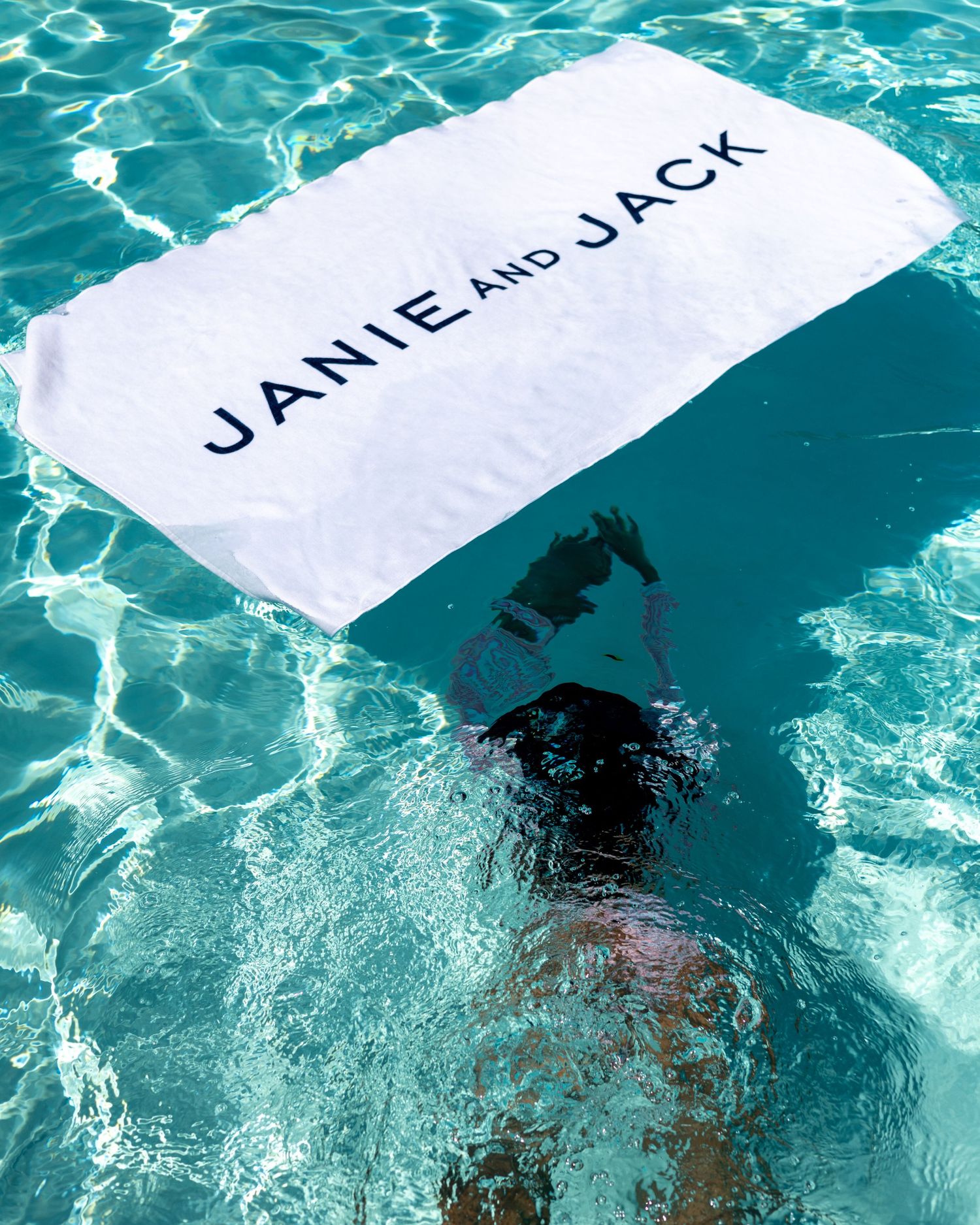 Person floating in a clear turquoise pool holding a white Janie and Jack branded towel.