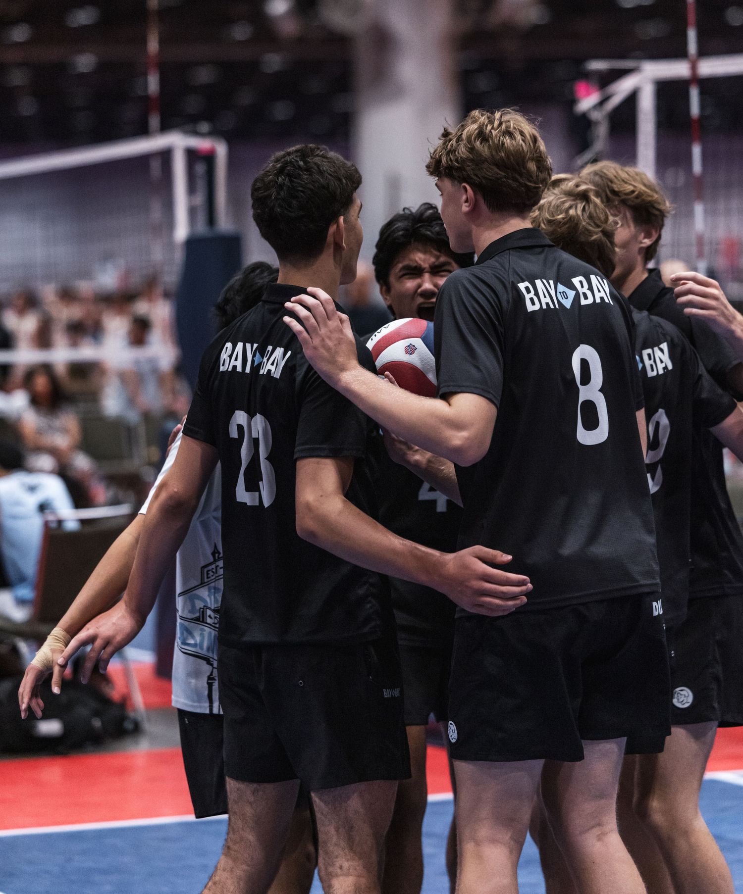 Youth volleyball players in black BAY-BAY jerseys huddle together on indoor court during competitive match.