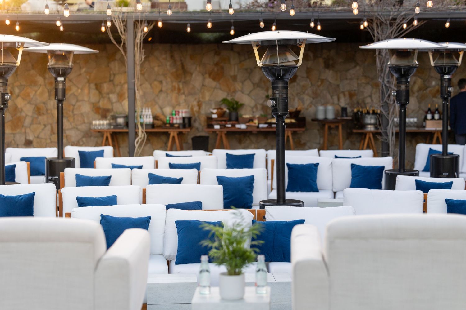 Elegant outdoor patio with white lounge furniture, blue pillows, patio heaters, string lights, and stone wall backdrop.