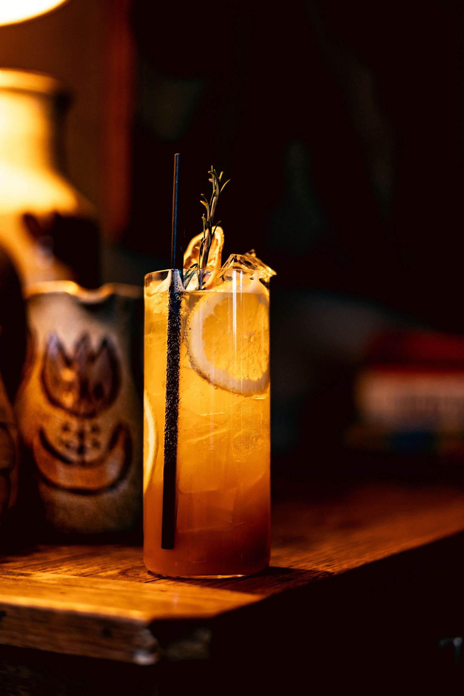 Amber cocktail with orange slice, rosemary, and black straw in a tall glass in warm bar lighting.