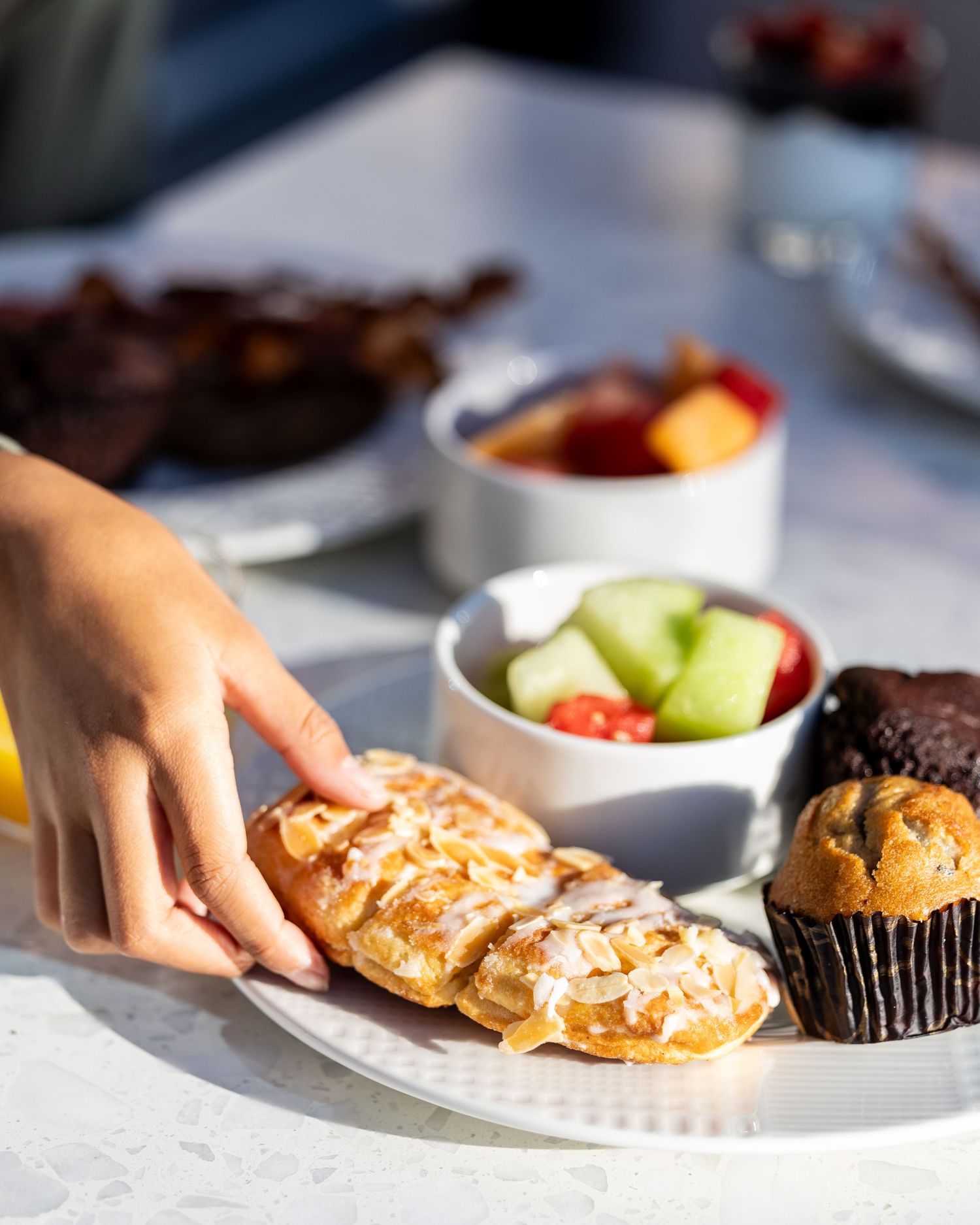 Hand reaching for pastry on plate with fruit and muffins.