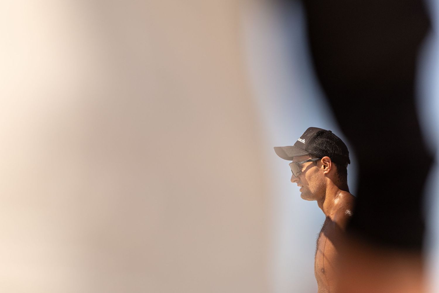 A shirtless man wearing a gray cap stands in profile, partially obscured by blurred foreground elements in soft light.