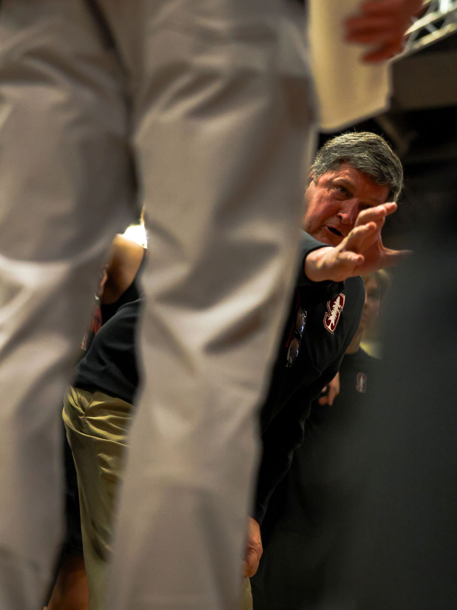 Coach gesturing instructions to players during a basketball game timeout, viewed between standing players.