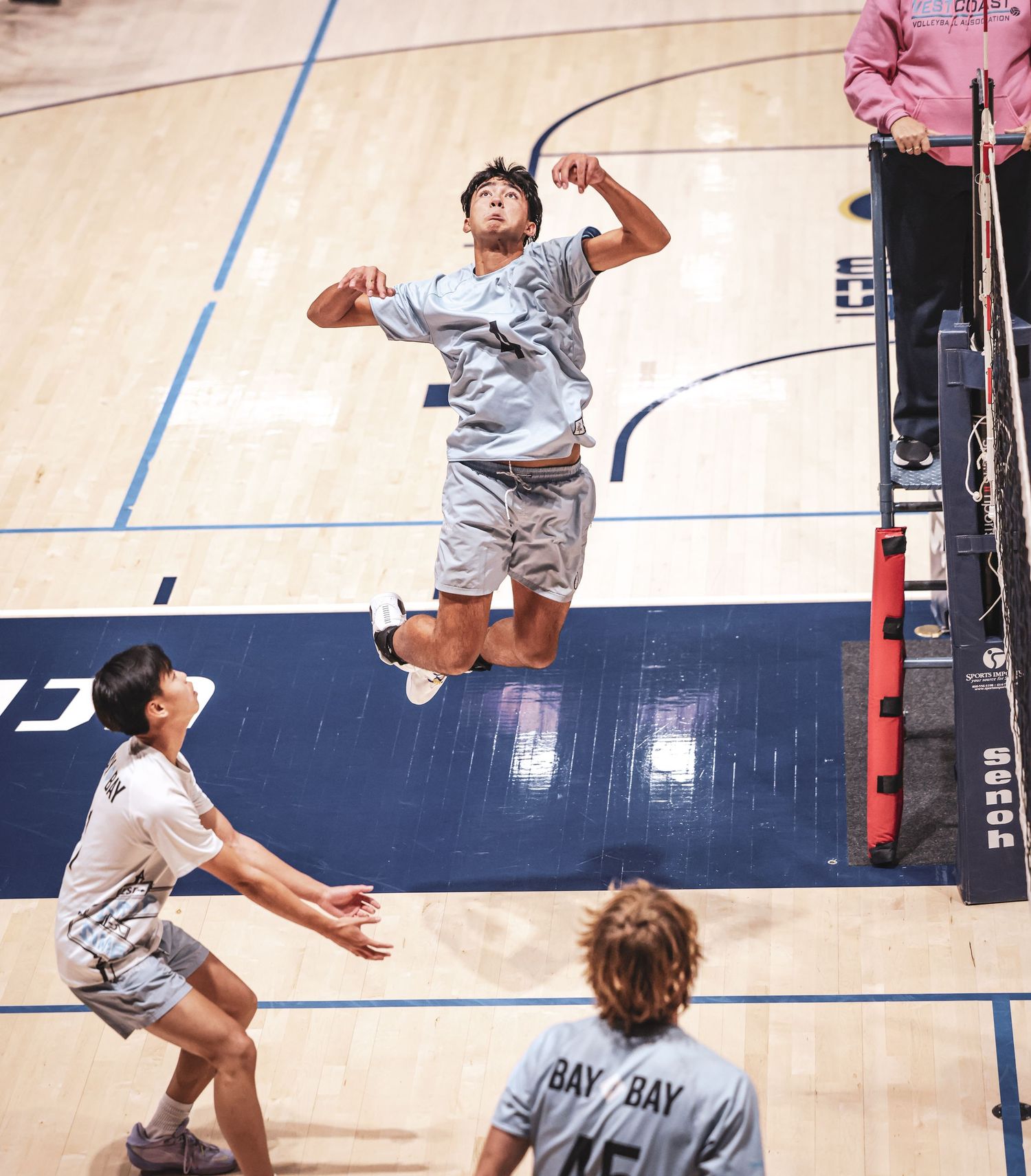 A volleyball player jumps to spike the ball during an indoor match, with teammates watching below.