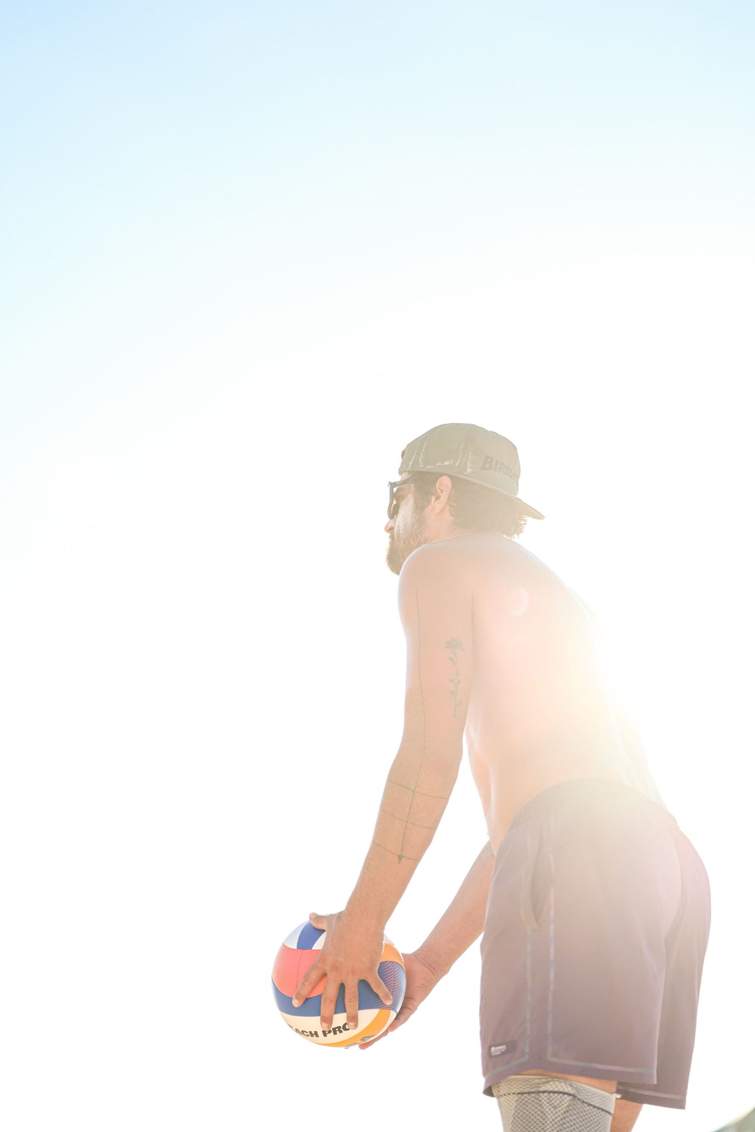 Shirtless man wearing a bucket hat and sunglasses holds a volleyball on a sunny beach.