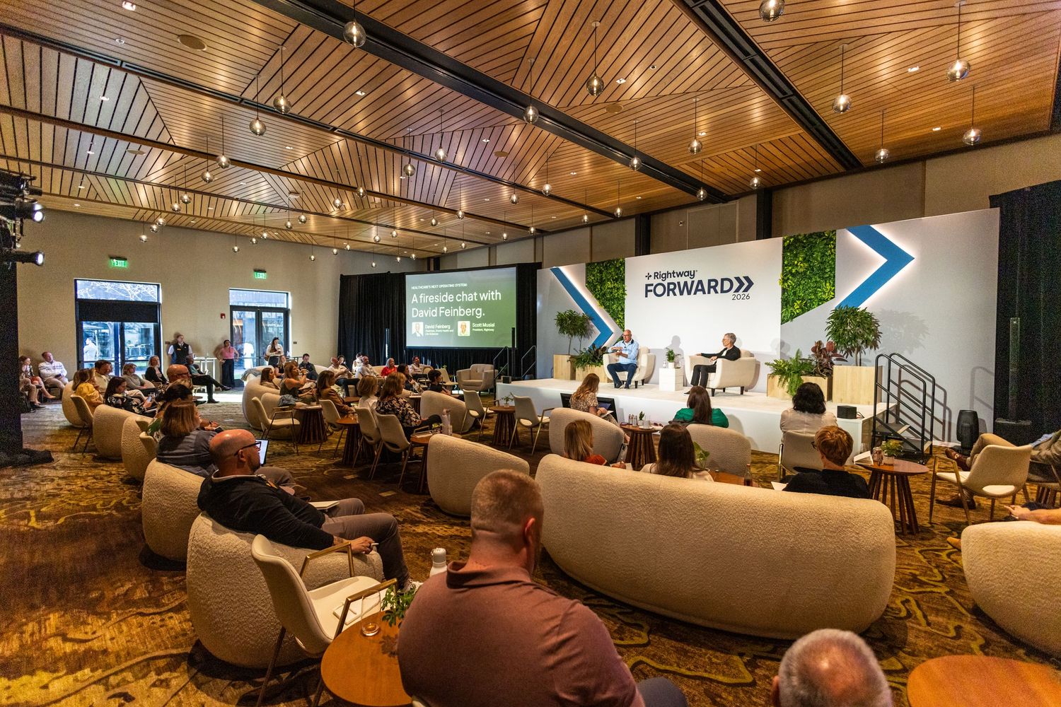 Conference attendees seated in a modern event hall facing a RightWay Forward branded stage and presentation screen.