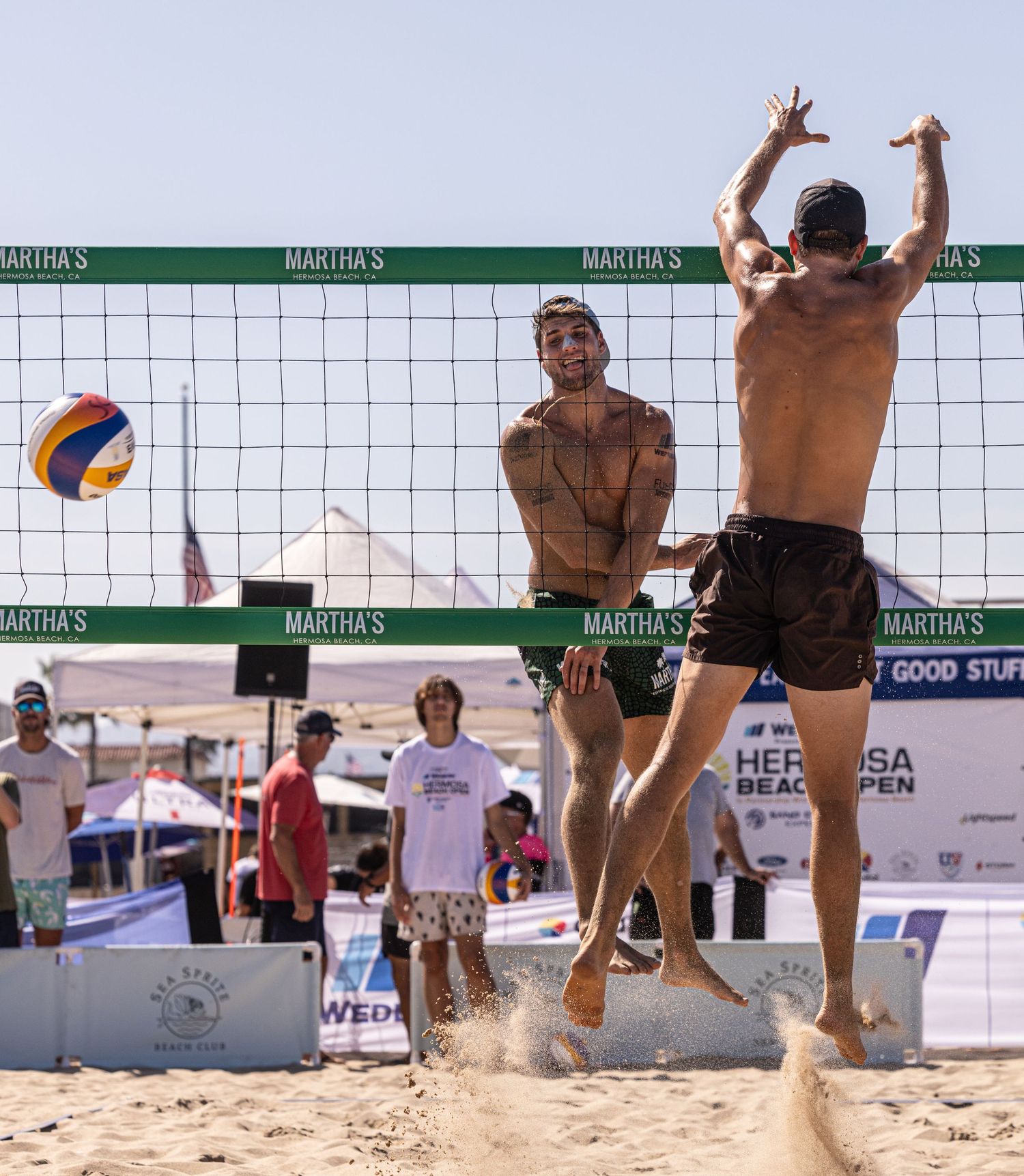 Two shirtless men play beach volleyball at Martha's Hermosa Beach Open, one jumping to spike the ball over the net.