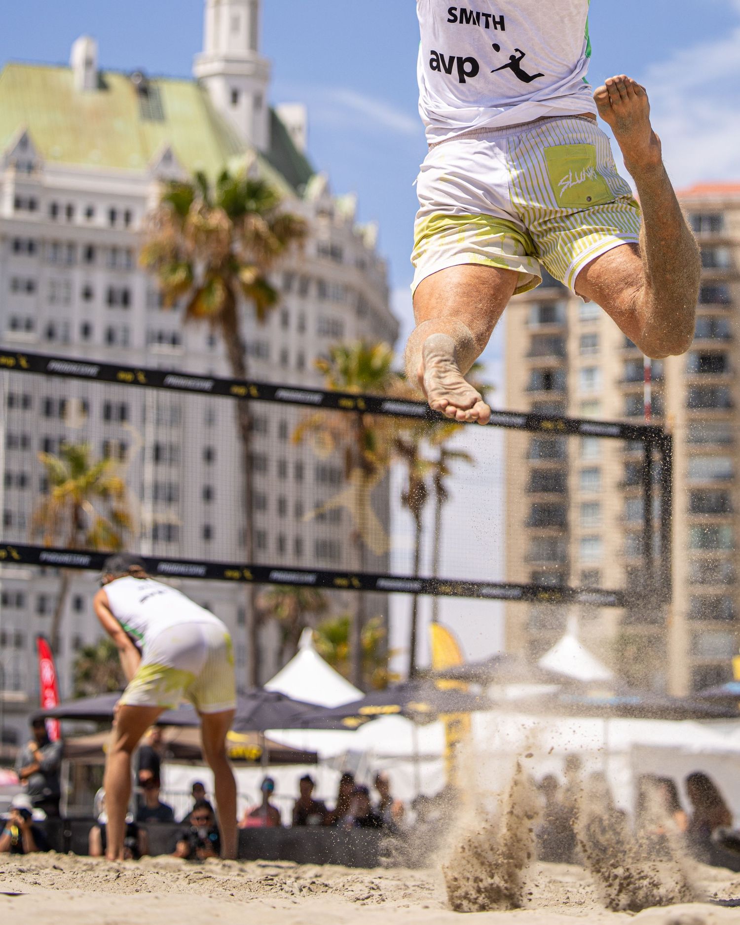 AVP beach volleyball player jumps high above the net during a professional tournament on a sunny beach.