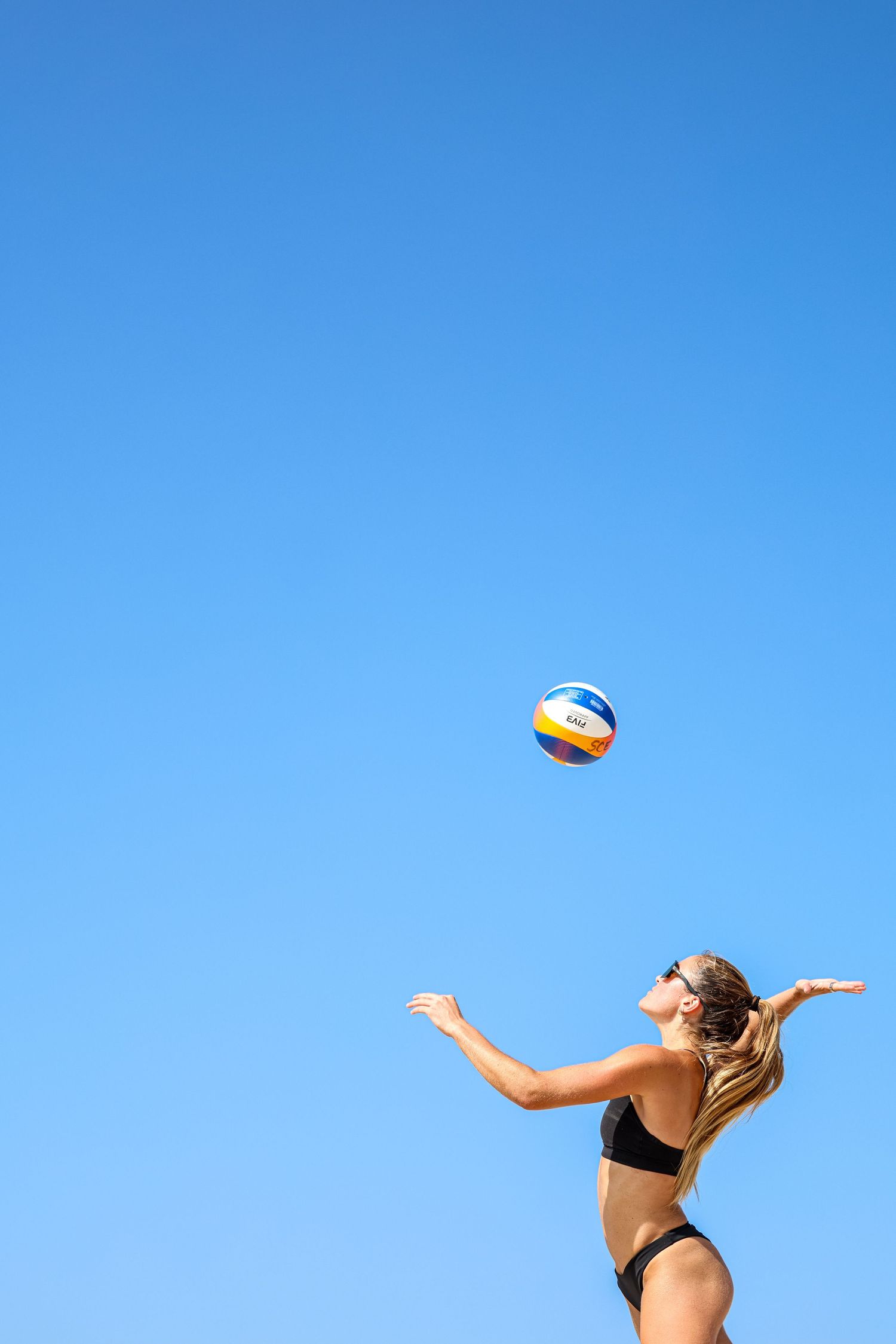 Woman in black bikini serving a volleyball against a clear blue sky on a sunny beach day.