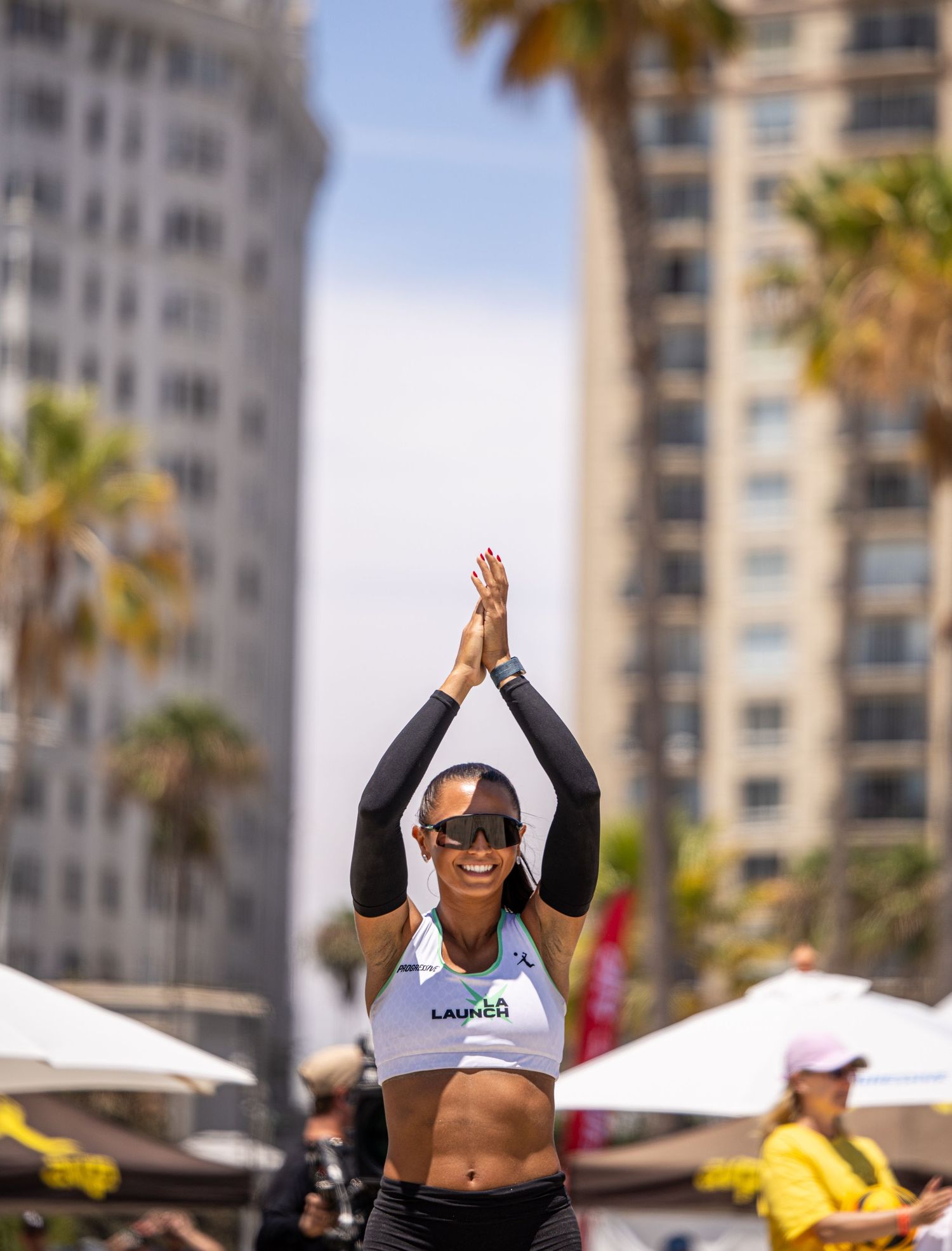 Athletic woman in white crop top and sunglasses claps overhead at outdoor beach volleyball event with palm trees and buildings.