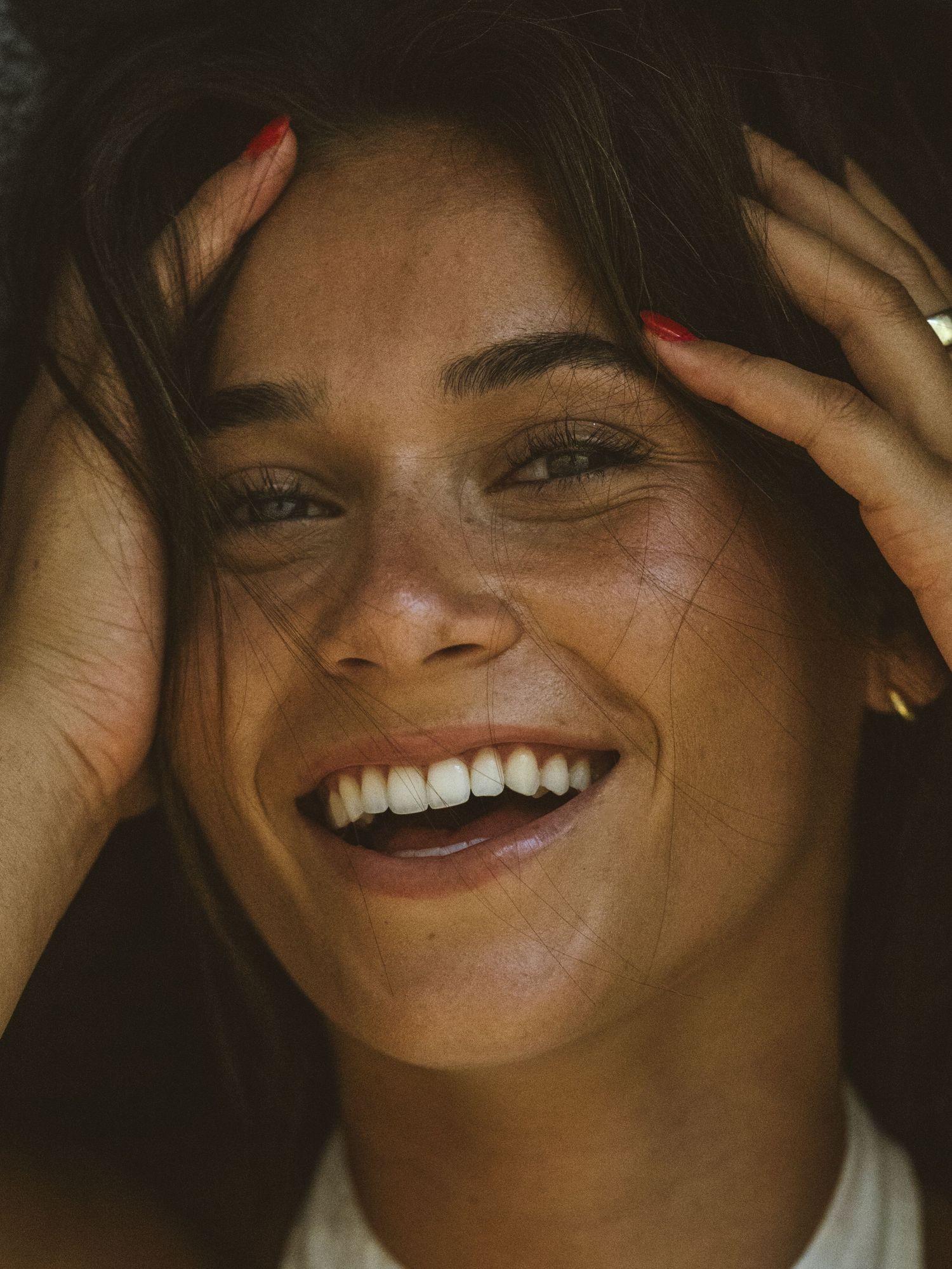 Smiling young woman with freckles, red nails, and gold ring holds her hair back, laughing joyfully in warm light.