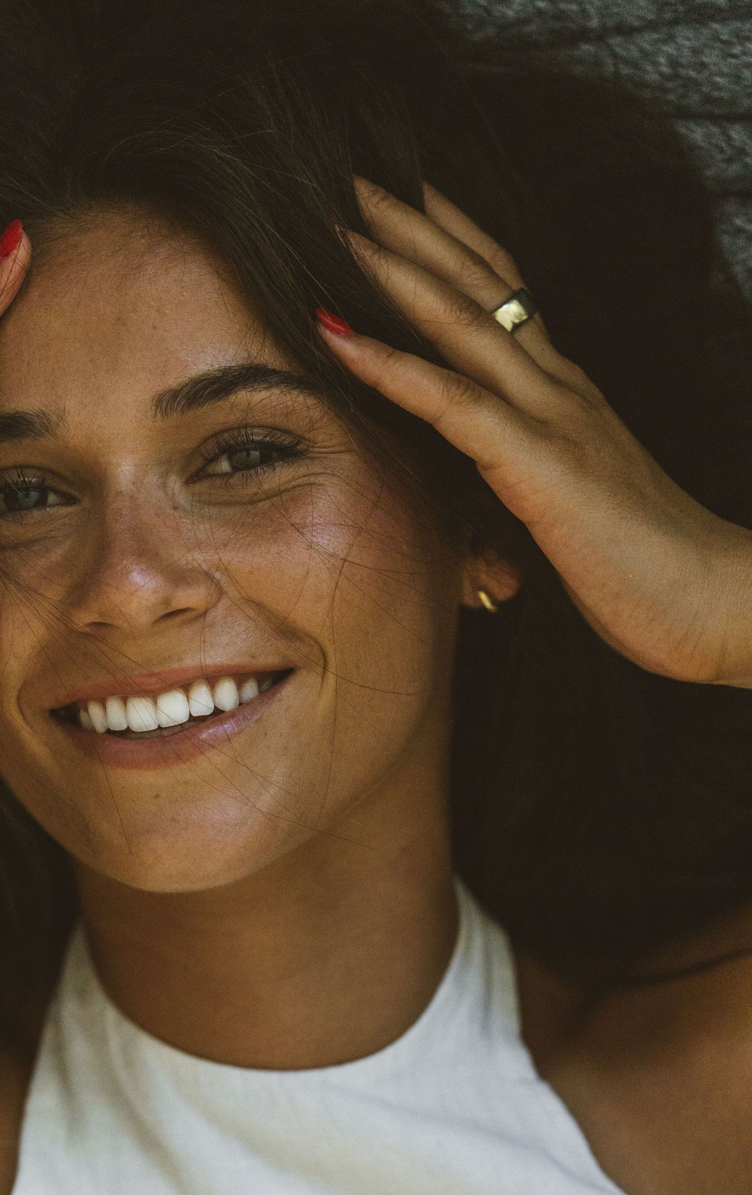 Smiling young woman with freckles, red nails, and a gold ring, posing with hand raised near her head outdoors.